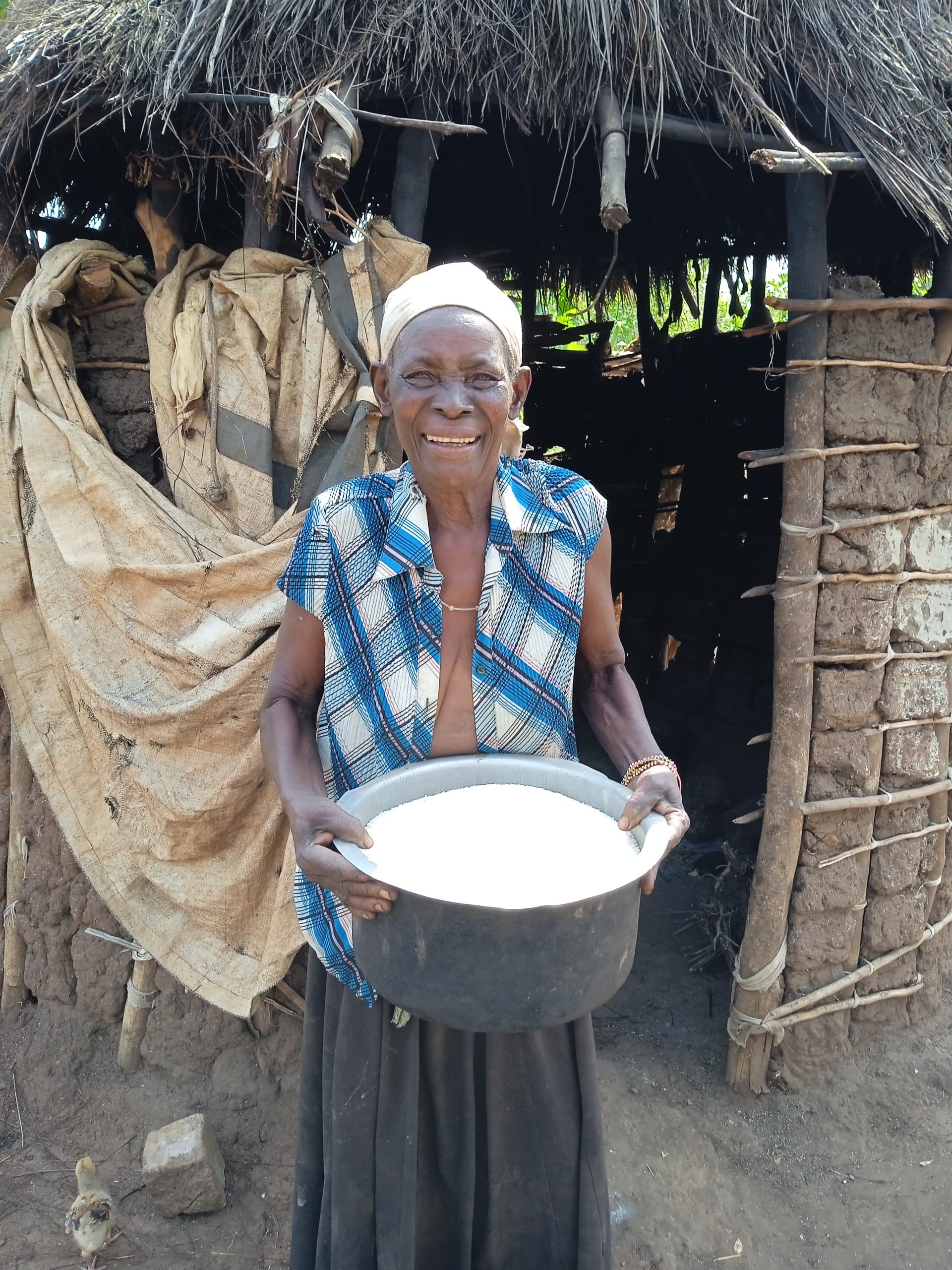 An elderly woman stands in her hut's doorway holding a black cooking pot.