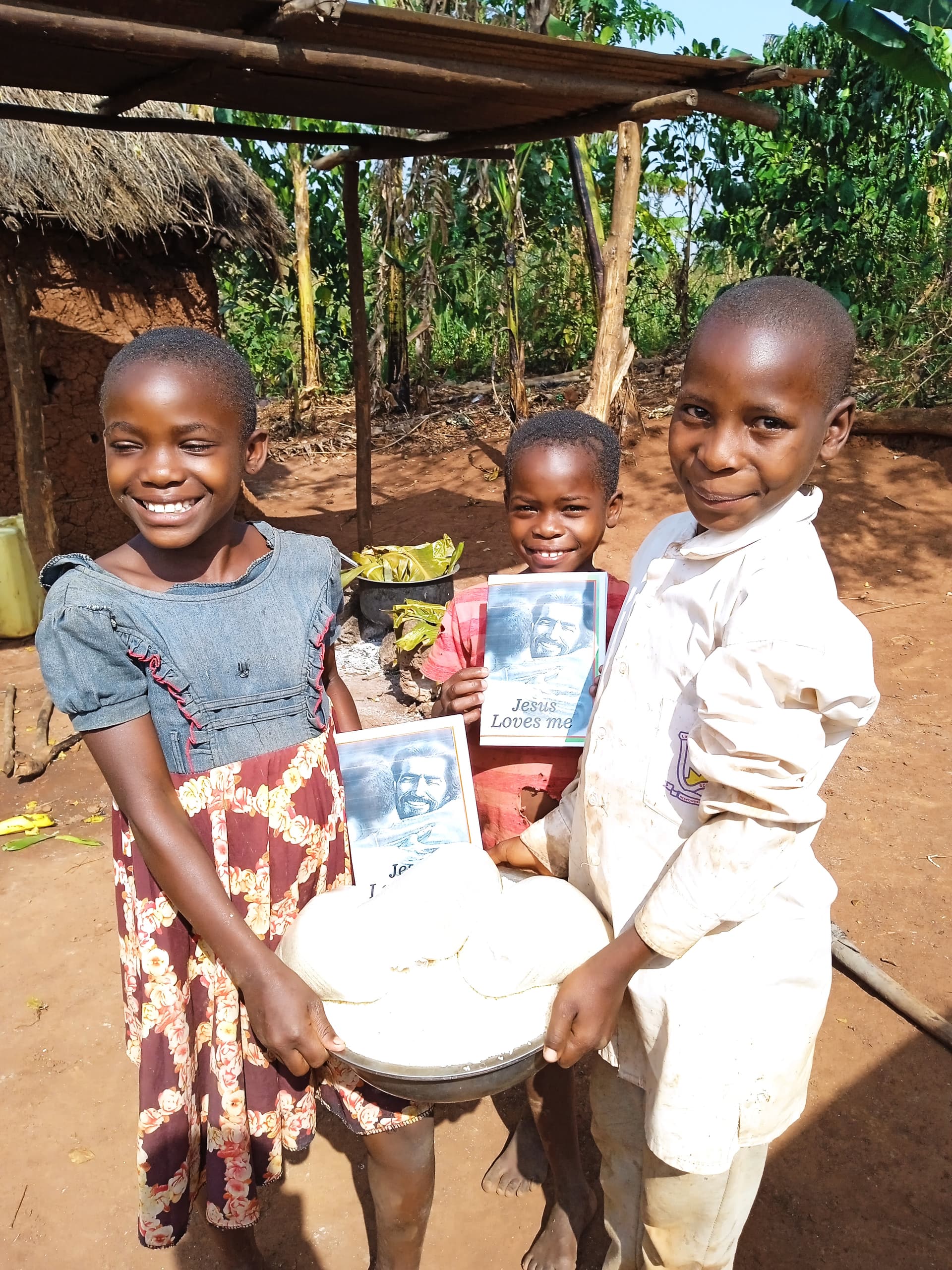 Three girls stand under a thatched shelter holding a basin with a gospel tract inside.