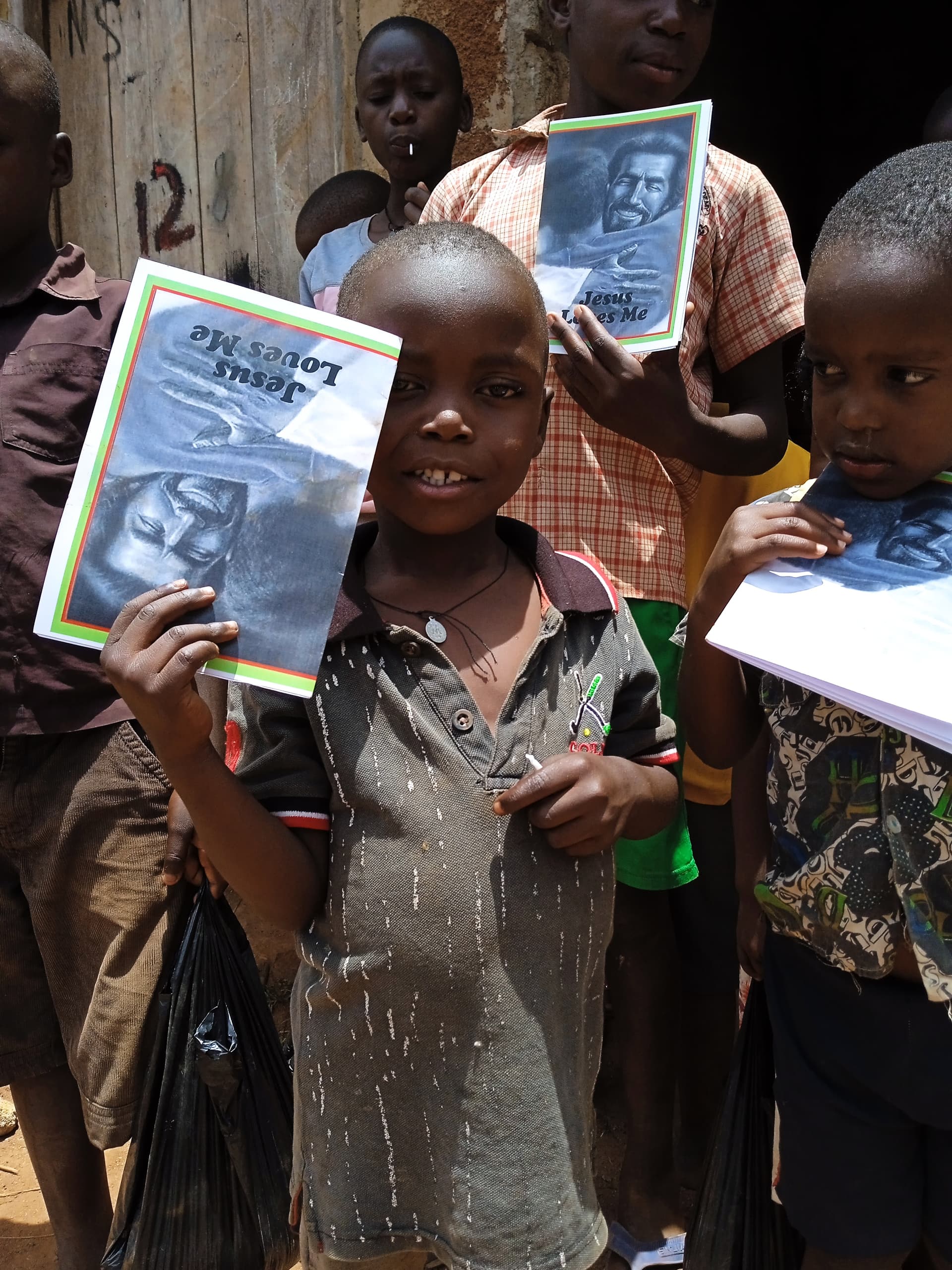 A child holds up a 'Jesus Loves Me' gospel tract booklet alongside other children.