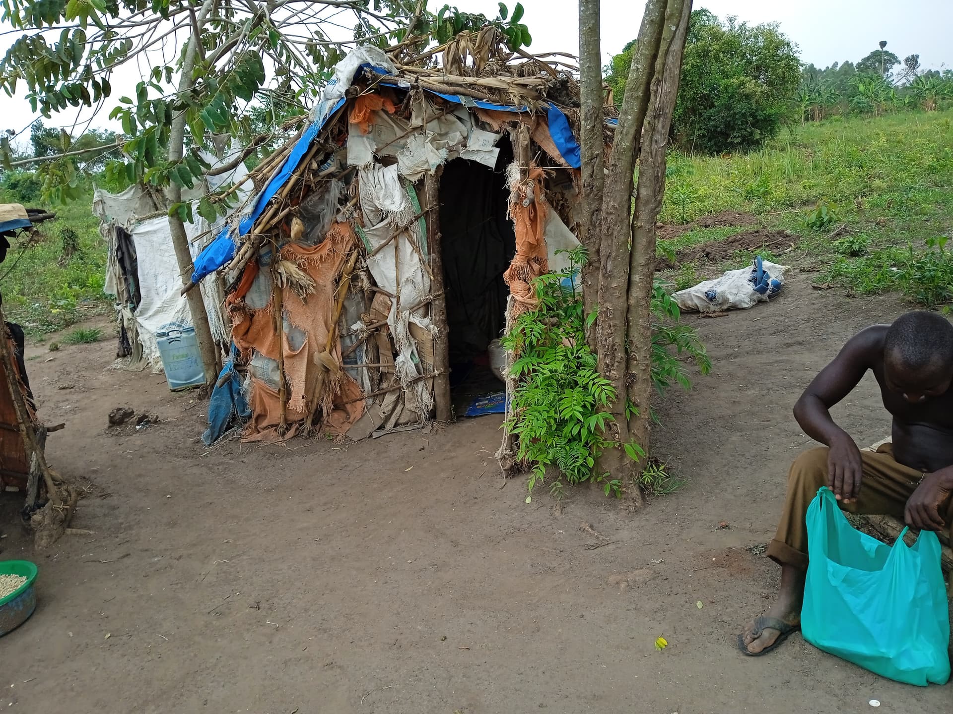 A mud-and-thatch shelter with a woman near the entrance and a teal bag on the ground.