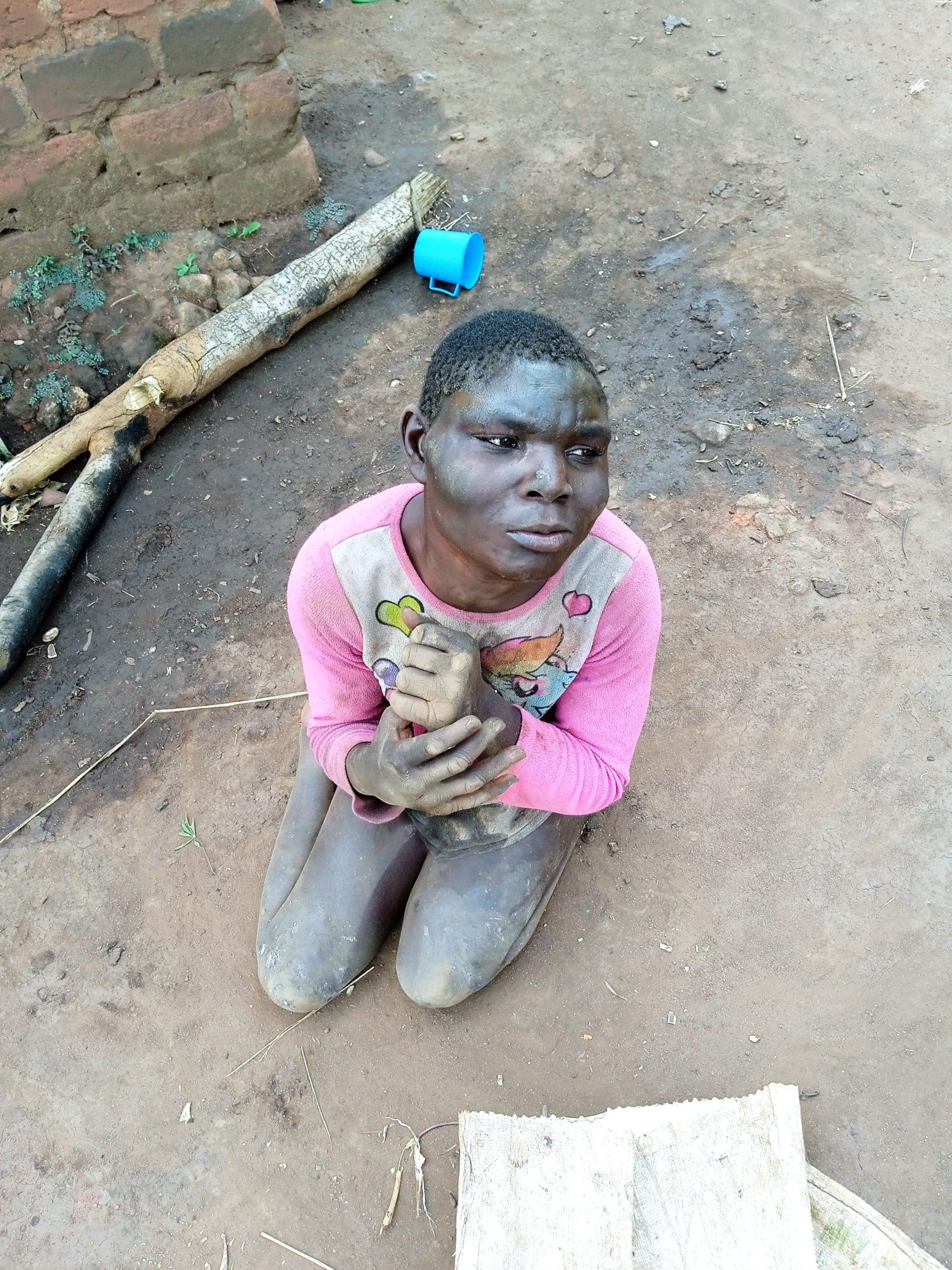 A boy with a physical disability kneels barefoot on bare dirt ground beside sticks and a small blue object.