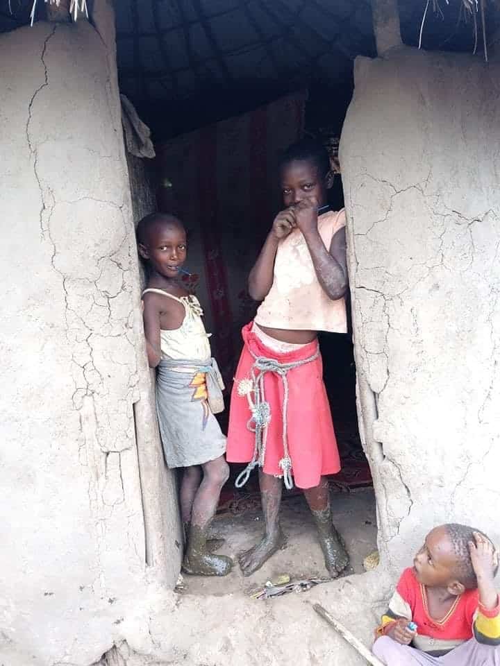 Two children and an infant stand at the doorway of a mud house.