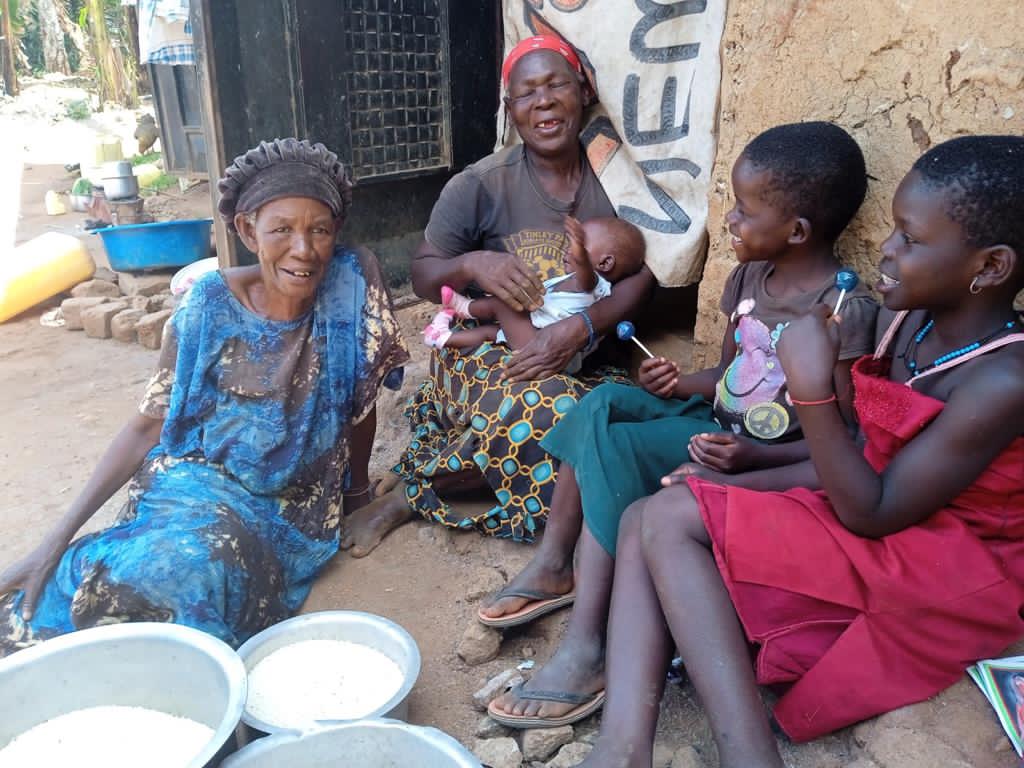 An elderly woman and children sit together outdoors near basins of food.