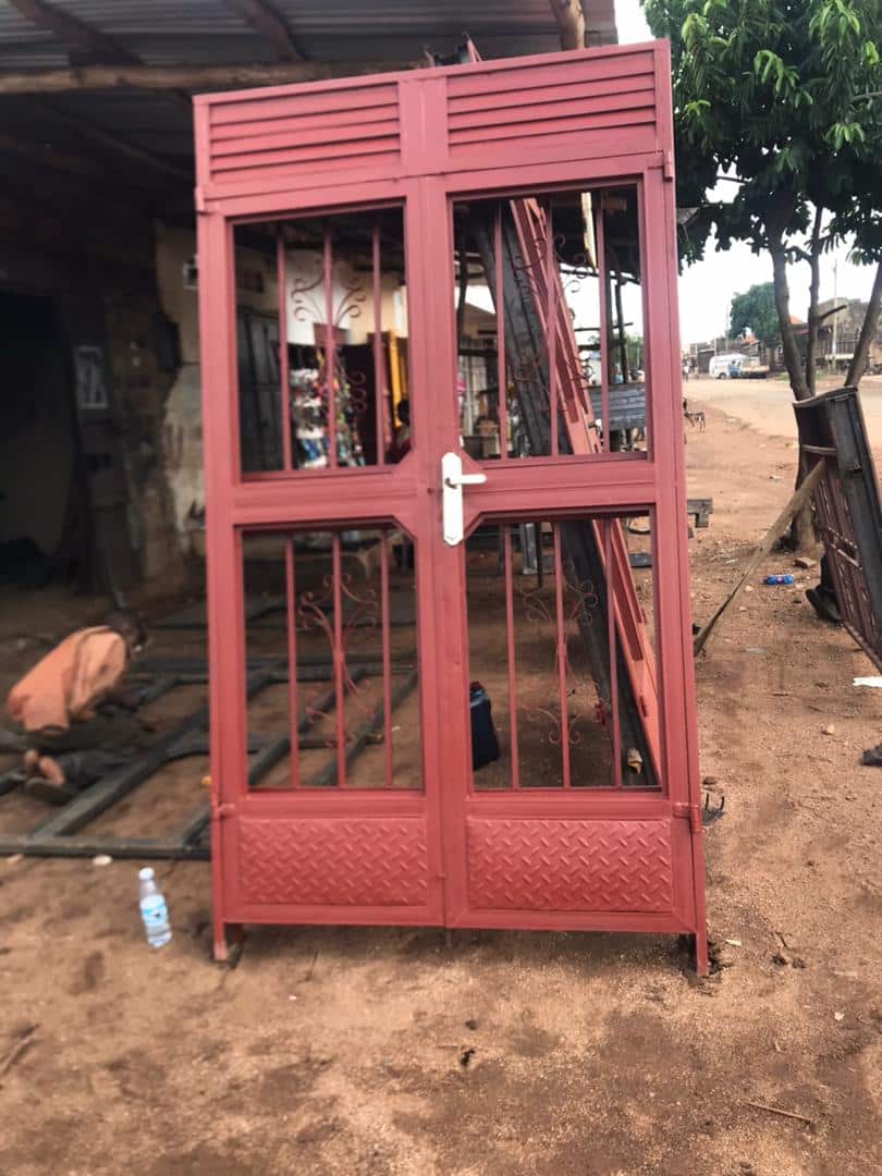 A red metal door frame with wire mesh security bars stands outside a building for Granny Angella's new house.