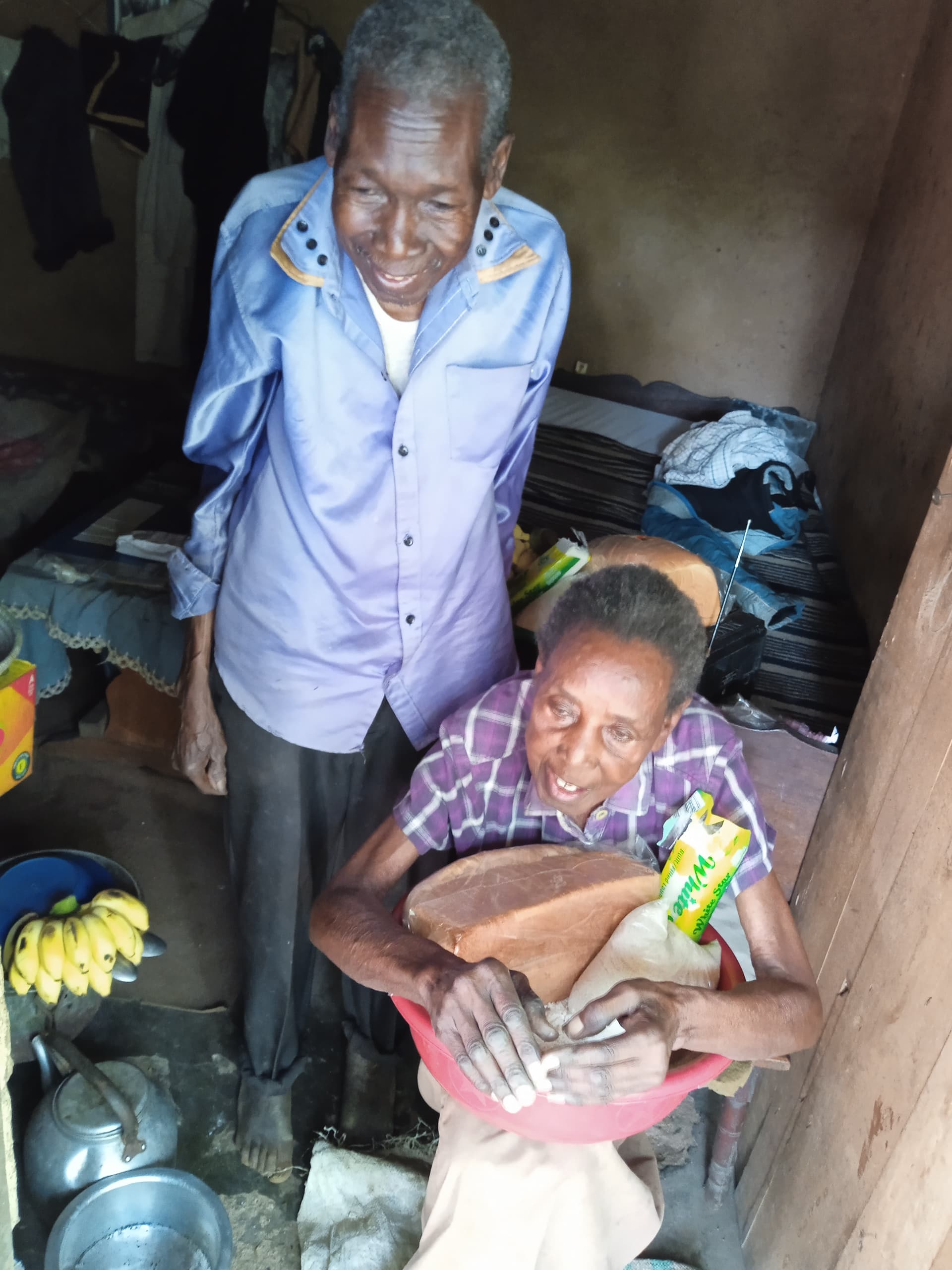 Elderly man in a blue shirt stands beside his wife, who sits holding a bowl of food items indoors.