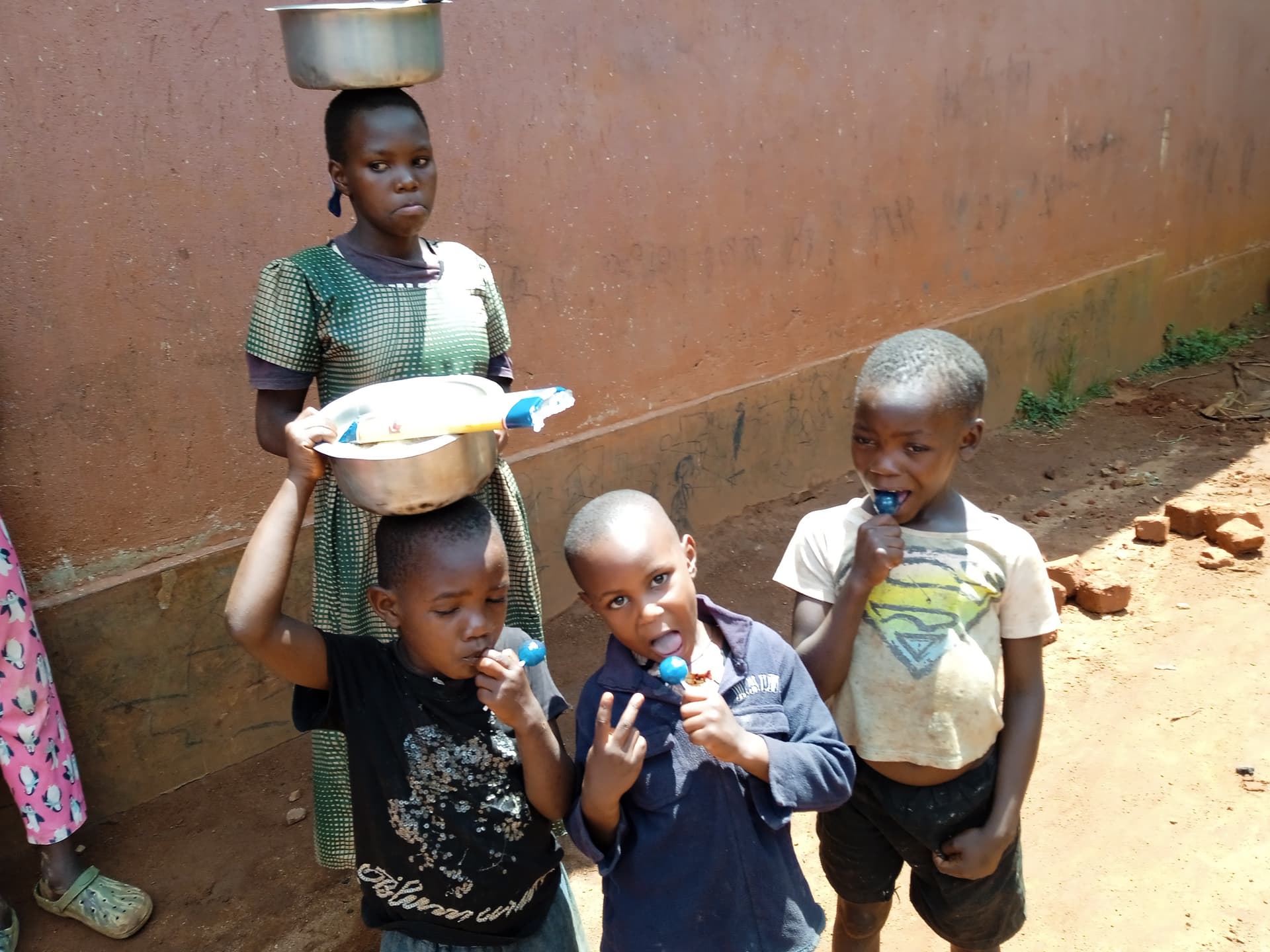 Group of Ugandan children balancing metal bowls on their heads outside a mud-brick wall.