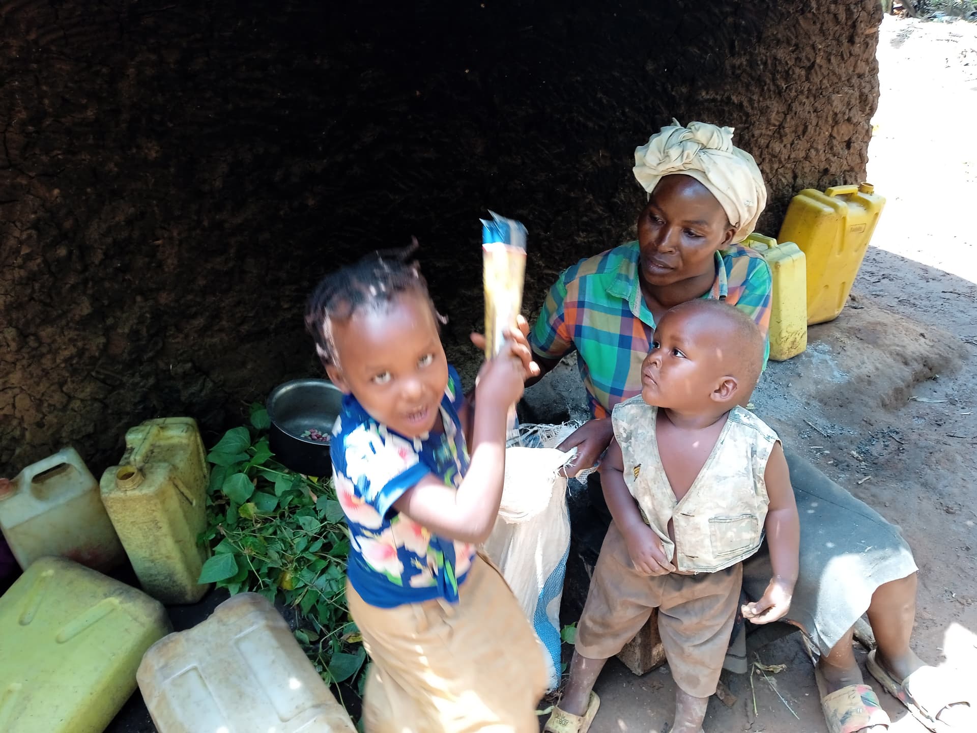 Two children sit under a tree, one holding a maize cob, with a woman and yellow jerrycans in the background.