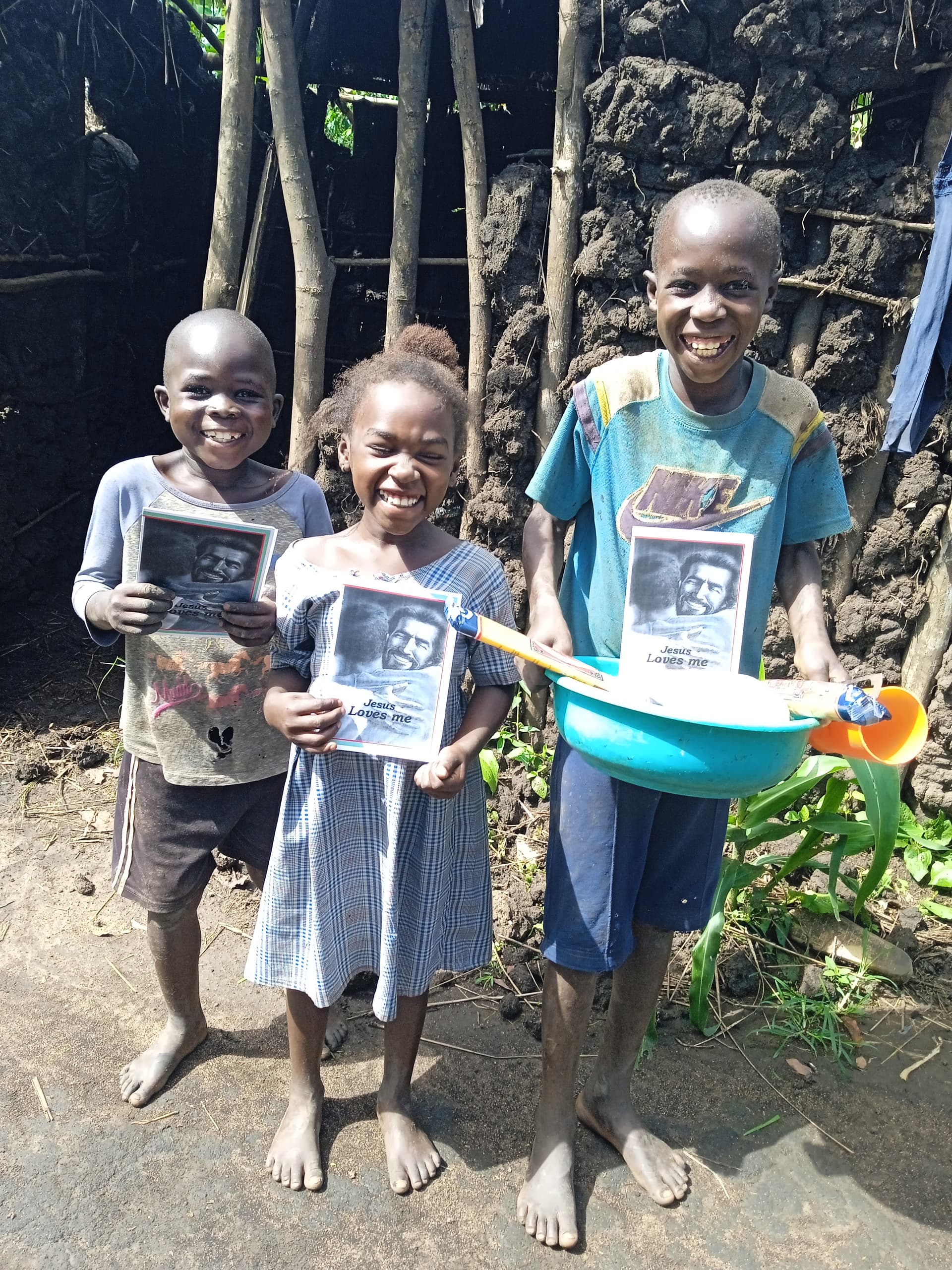A group of children in matching outfits stand by a stone wall holding photographs and a blue basin.