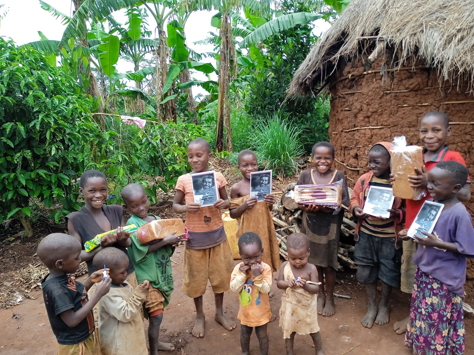A group of children stand outside a mud house holding photographs and food containers.