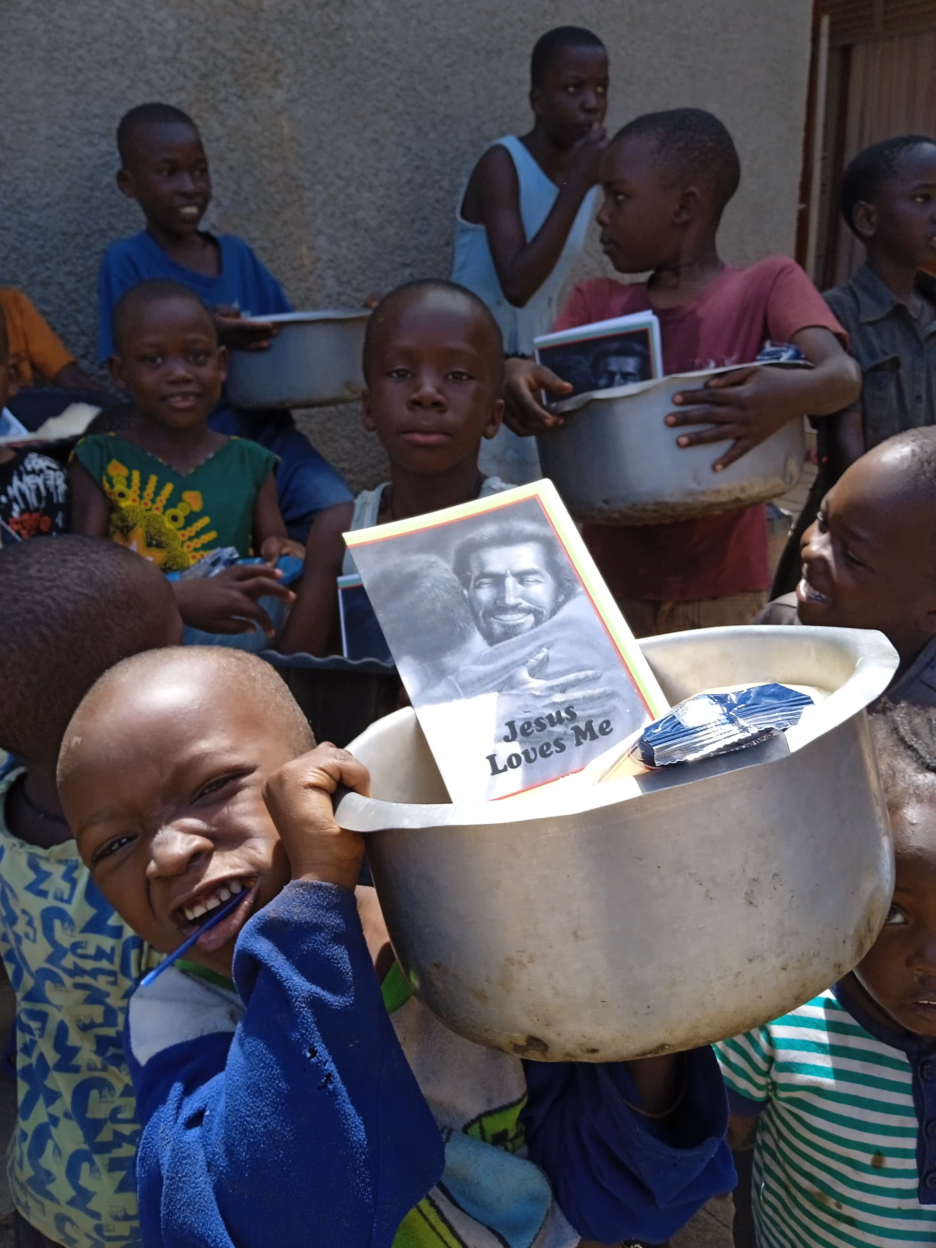A boy holds a large metal pot containing a photograph and a packaged food item, with children gathered behind him.