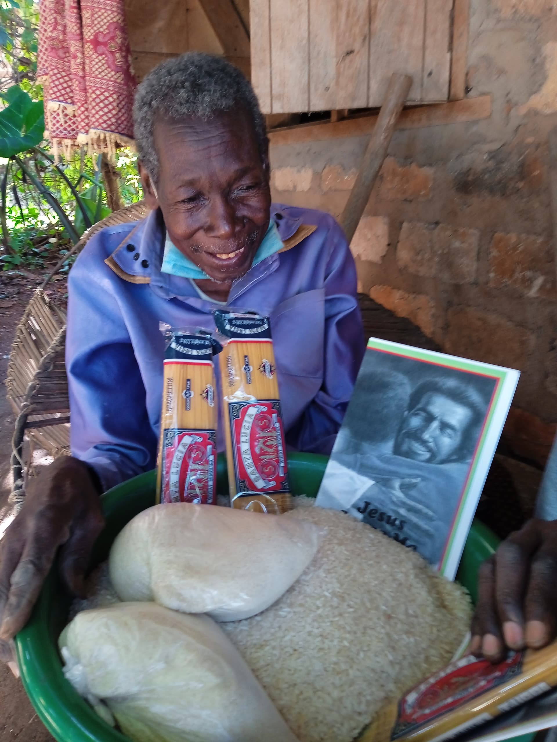 An elderly woman smiles while holding a basin with bread and food items and a photograph.