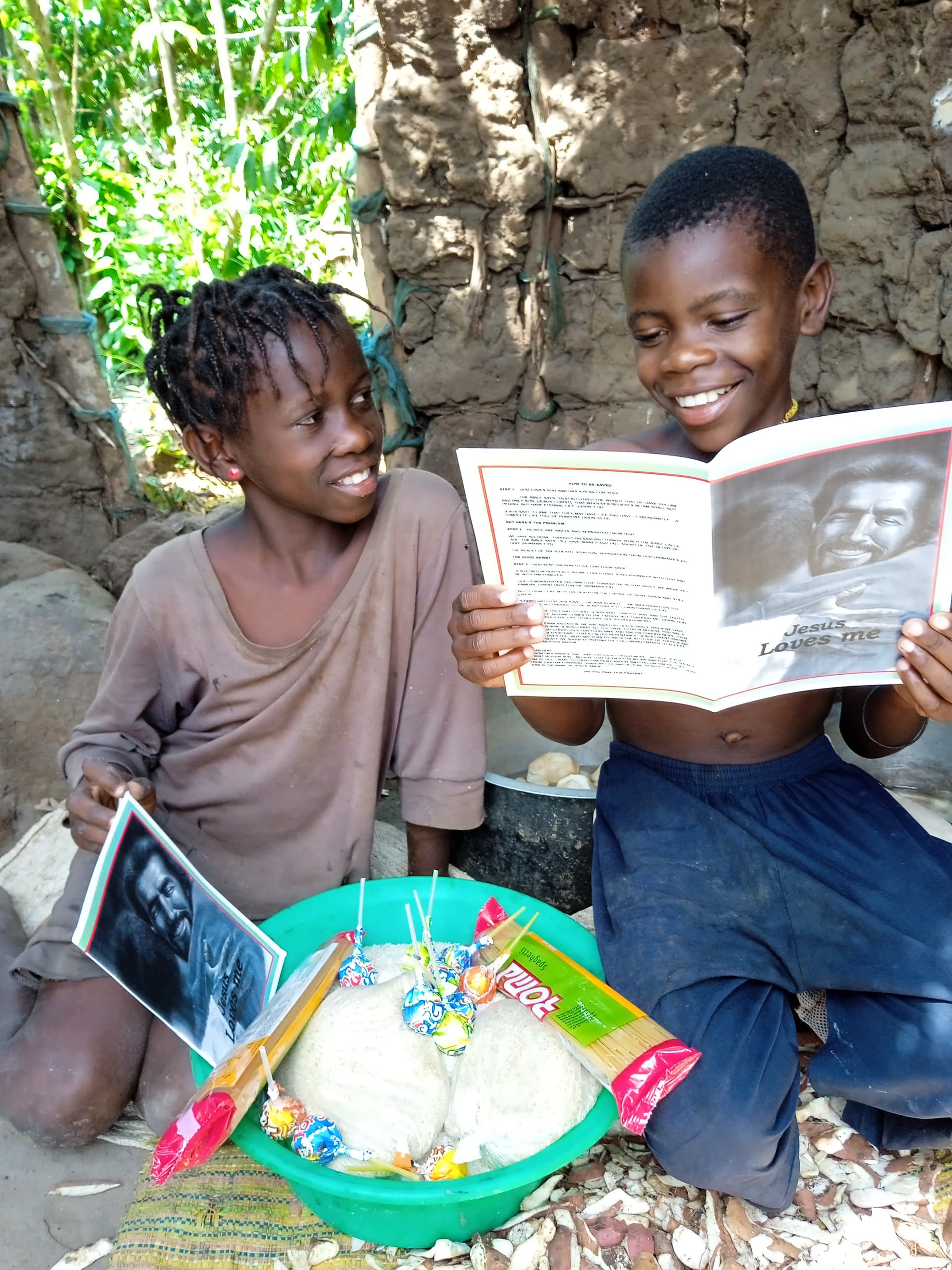 Two smiling children sit together outdoors, a boy holding a photo booklet beside a colorful basin of food items.