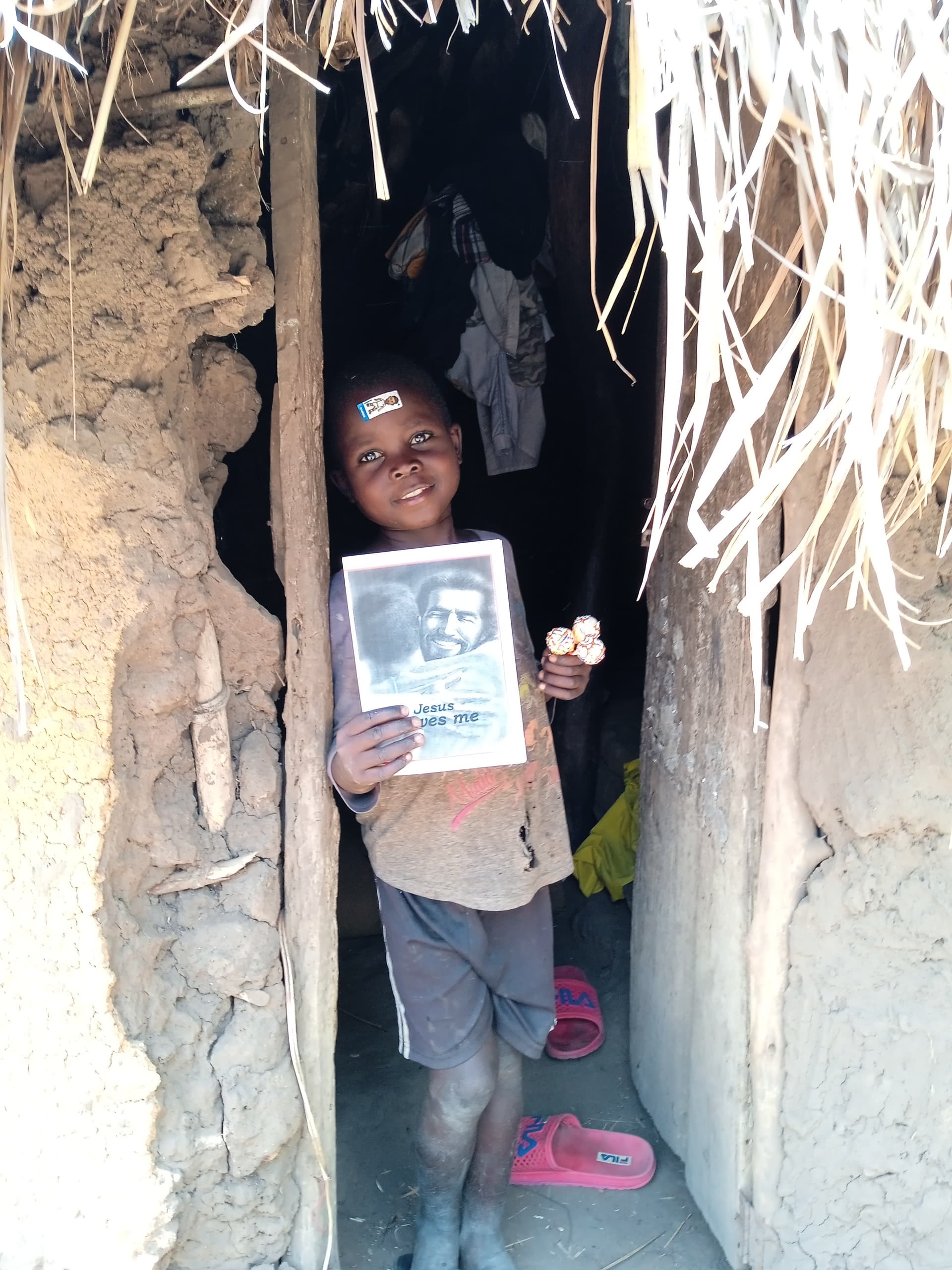A boy stands in the doorway of a mud house holding a photograph and a booklet.