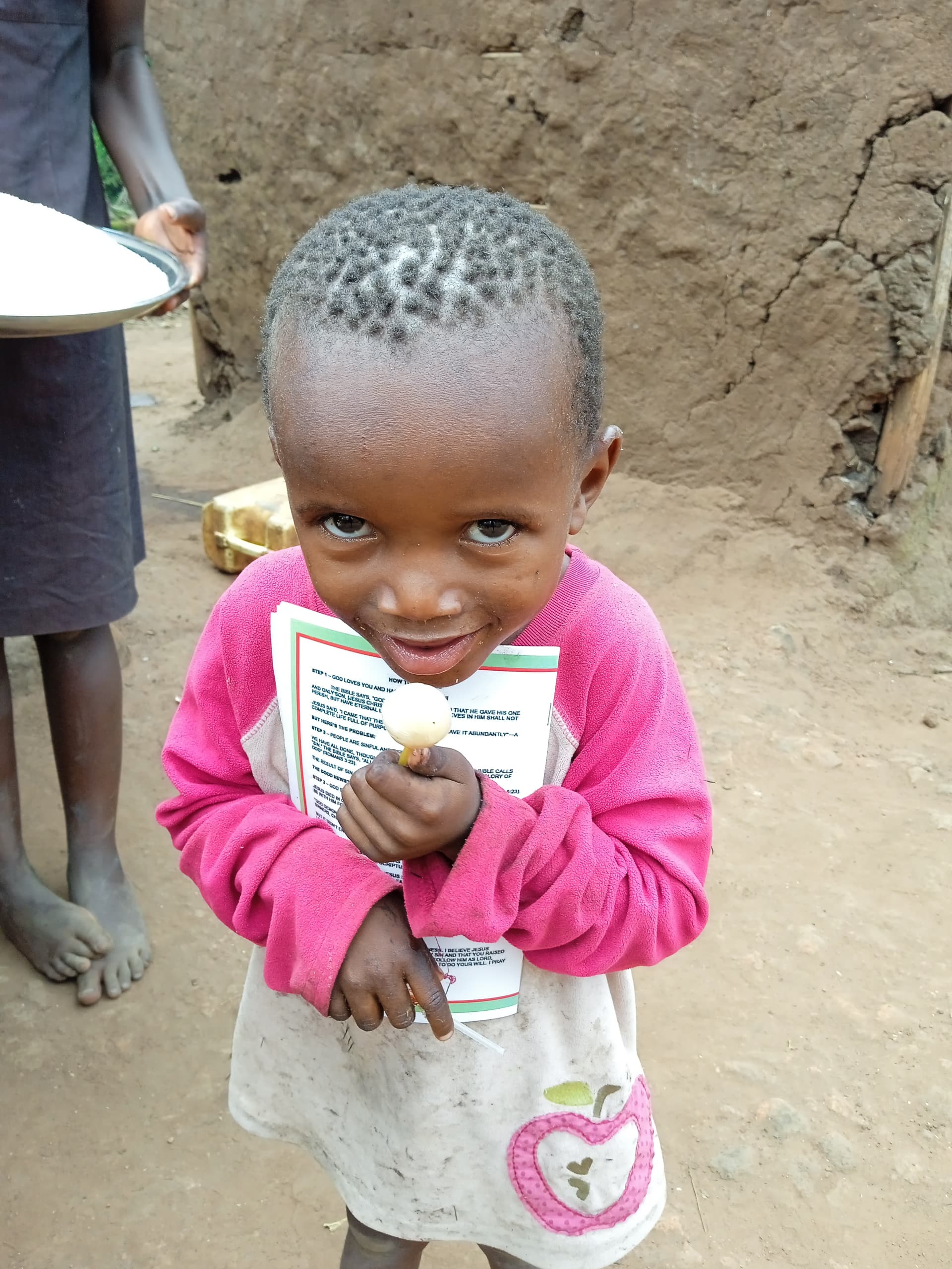 A young child in a pink shirt stands outside a mud house holding a photograph and a book.