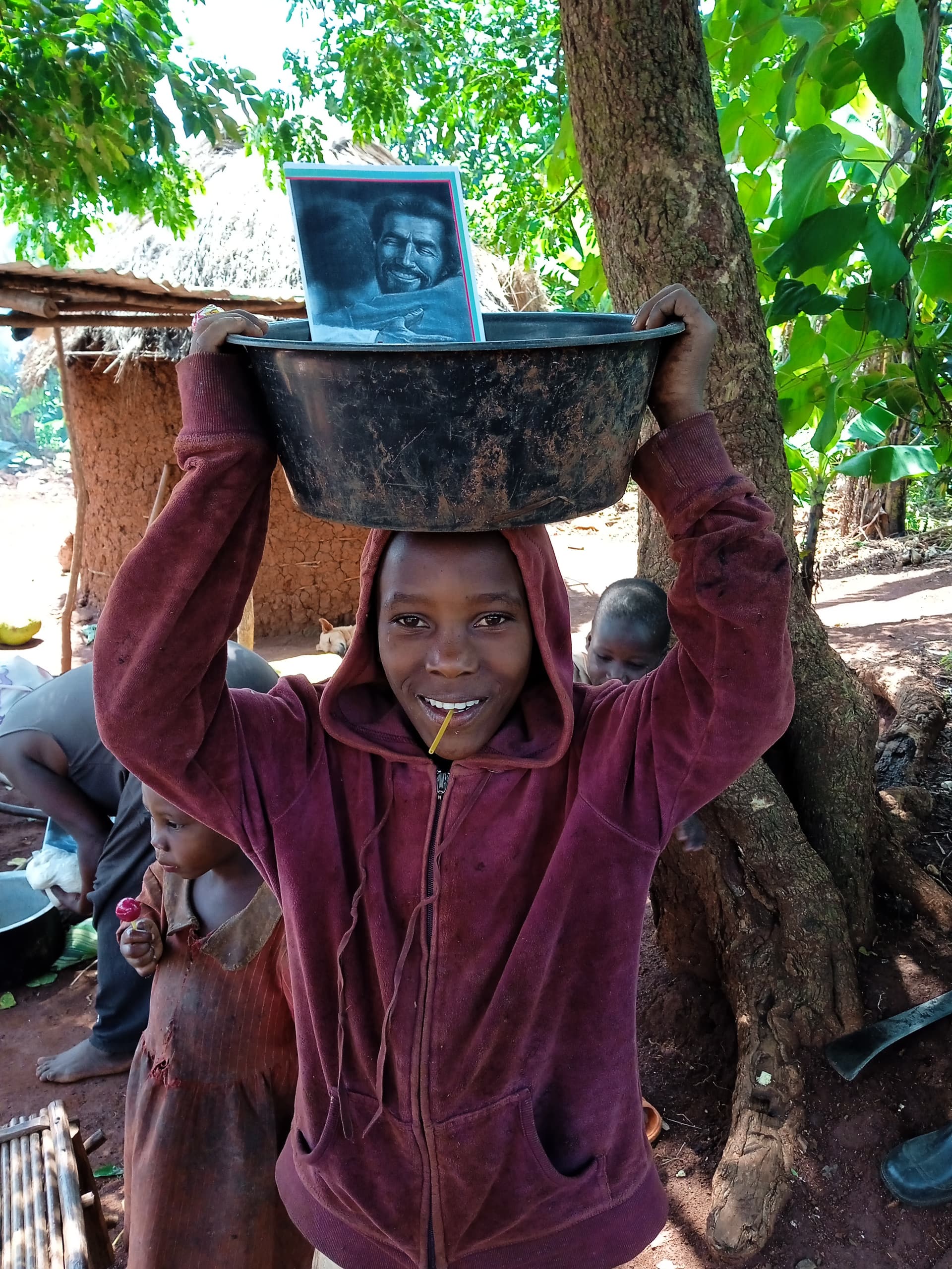 A girl balances a metal pot holding a photograph on her head while a boy in a maroon jacket stands nearby.