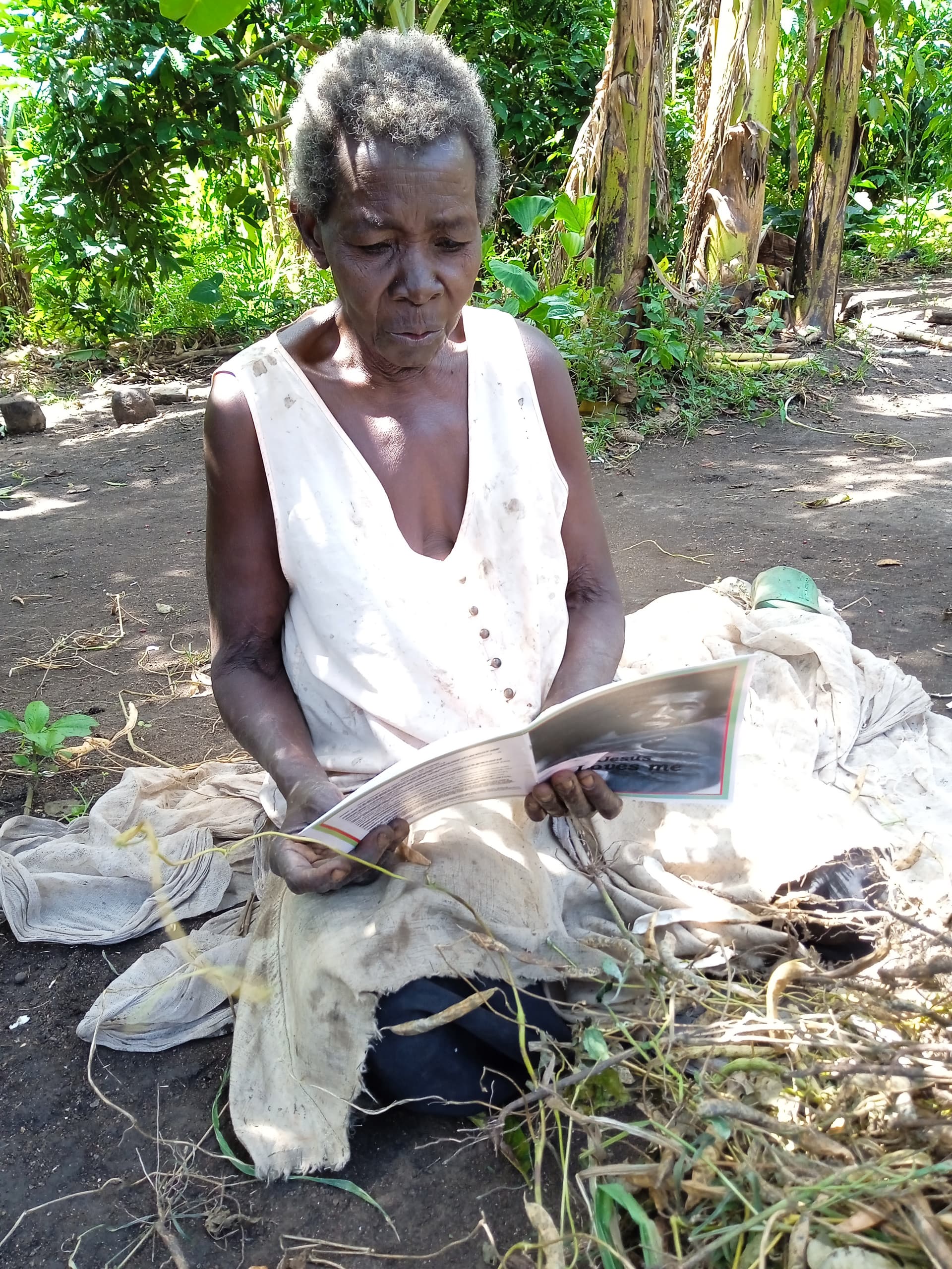 Elderly woman sits outdoors peeling cassava by hand near a cooking area.