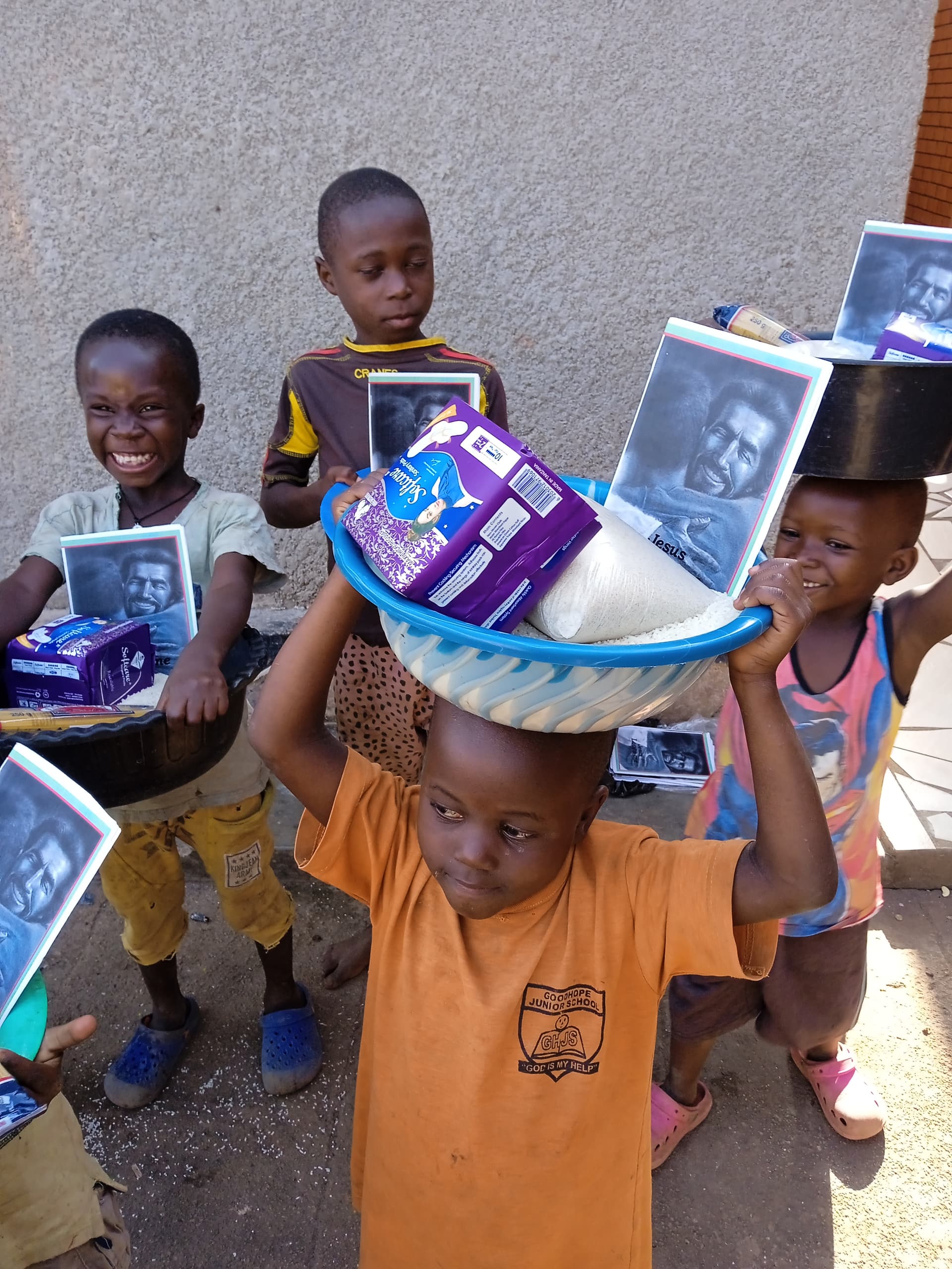 Boy in an orange shirt carries a basin of food supplies and a photo on his head, other children around.