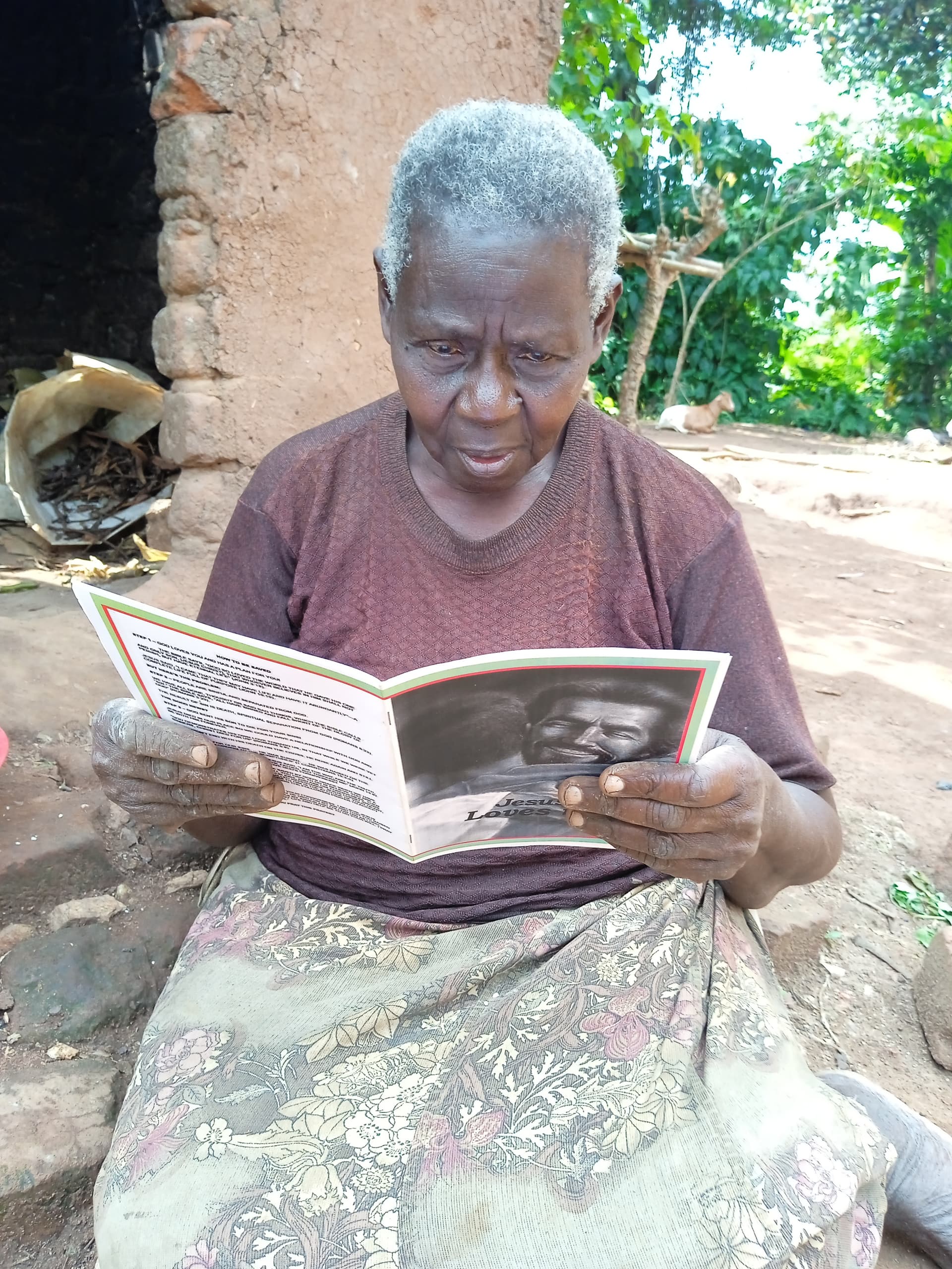 Elderly woman with gray hair sits outdoors reading an open booklet against a mud wall.