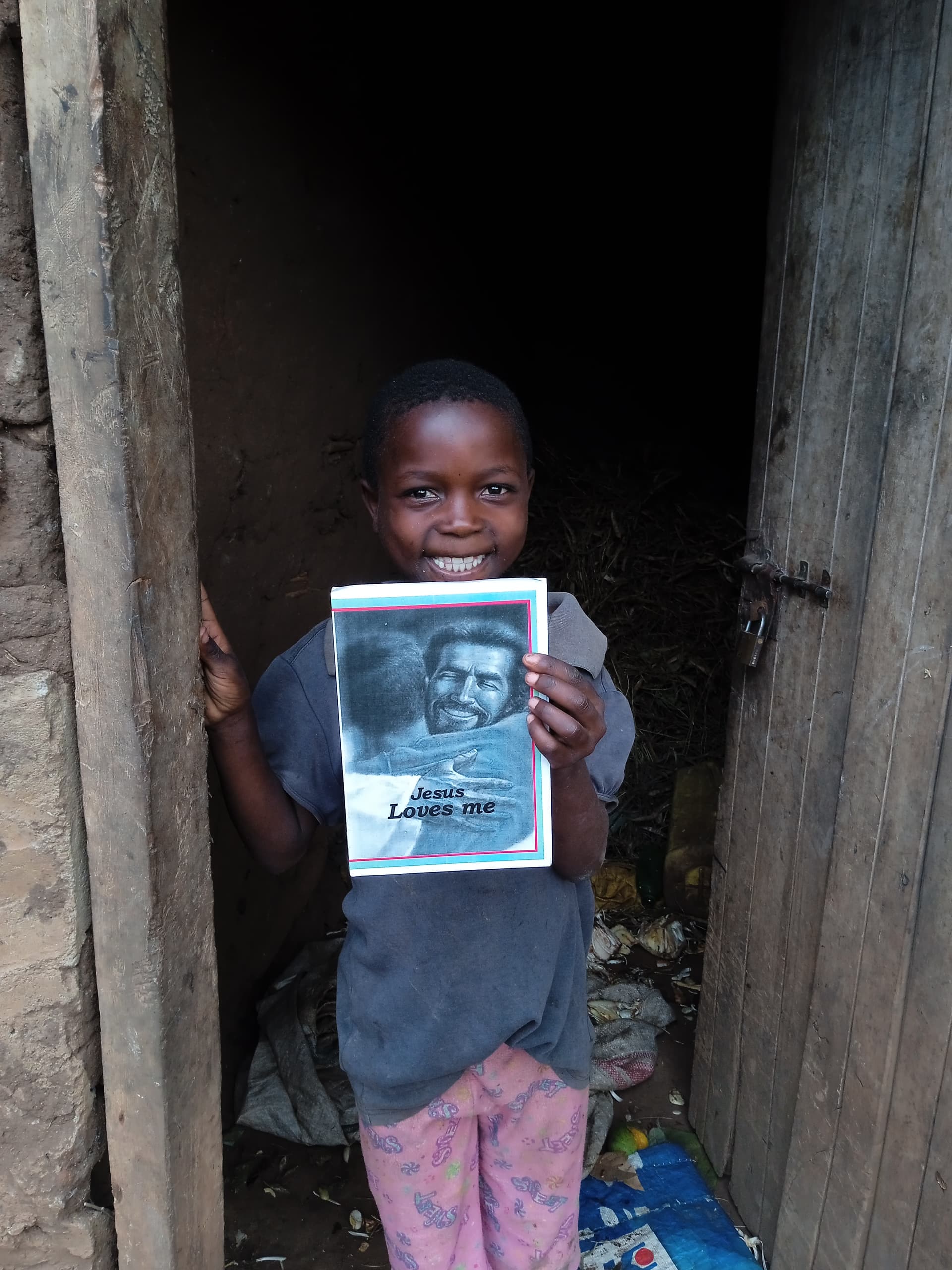 Boy standing in the doorway of a mud home, holding a gospel tract booklet.