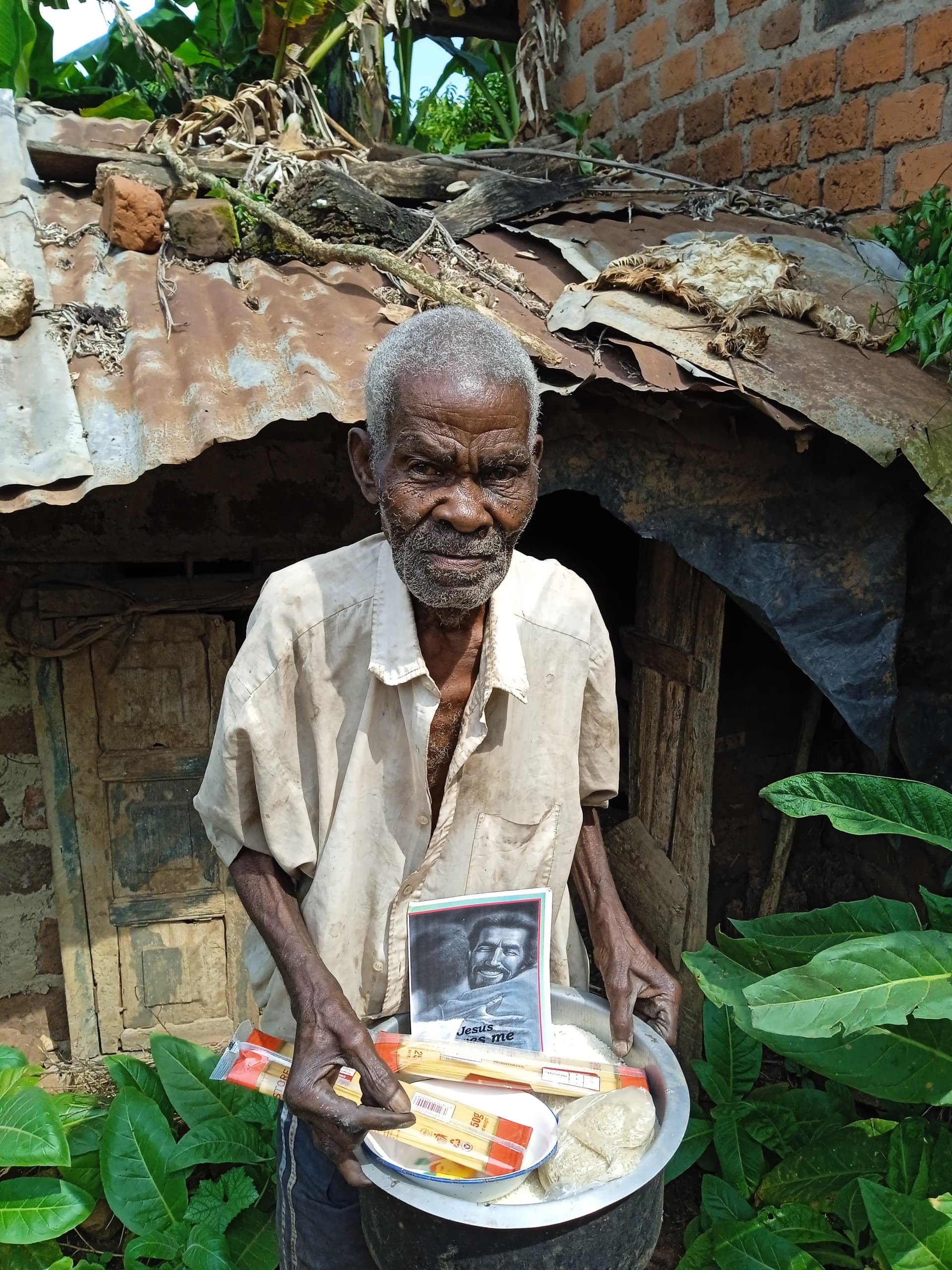 Elderly man stands under a thatched shelter holding a basin of food supplies and a photo booklet.
