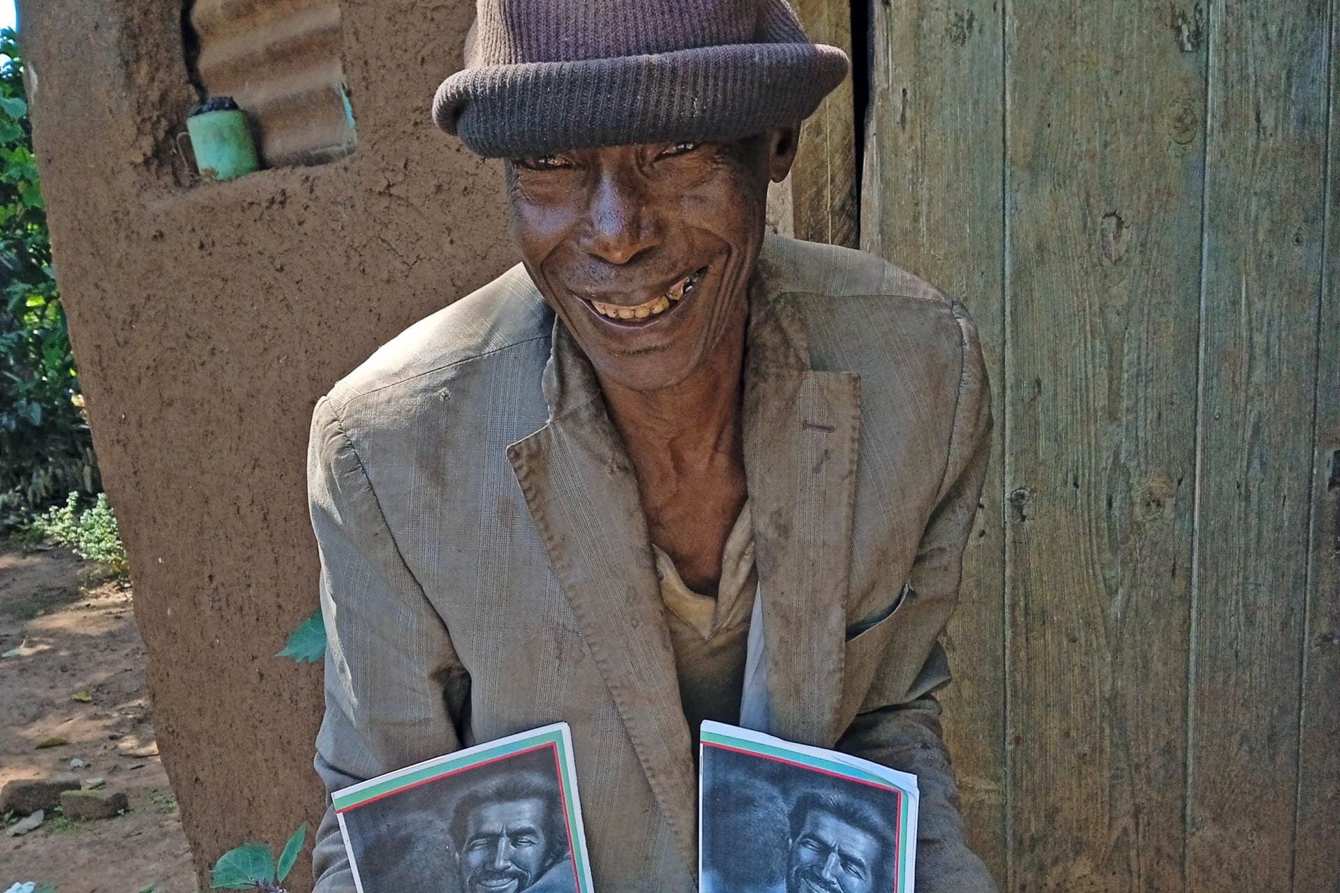 Elderly man in a hat and coat holds a basin with two photo booklets near a wooden wall.