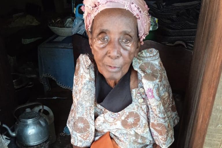 Elderly woman in a pink headscarf and patterned dress sits indoors with hands clasped.