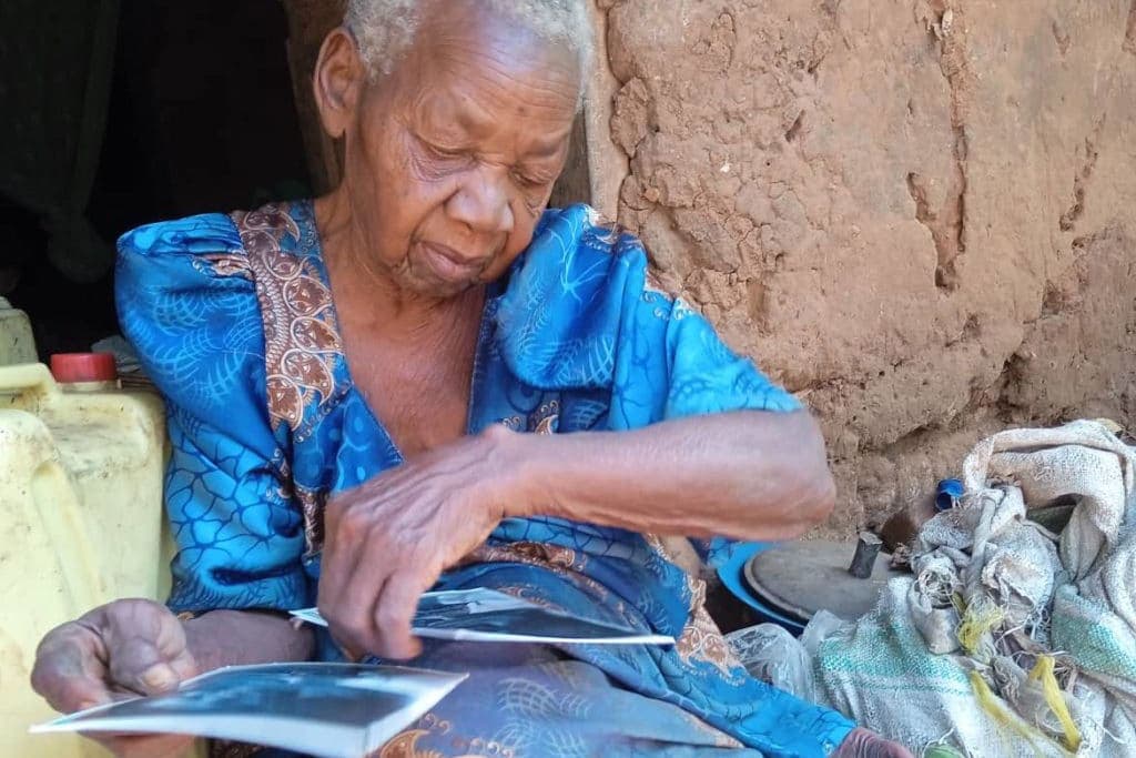 Elderly woman in blue clothing sits outdoors sorting through papers beside sacks of food.