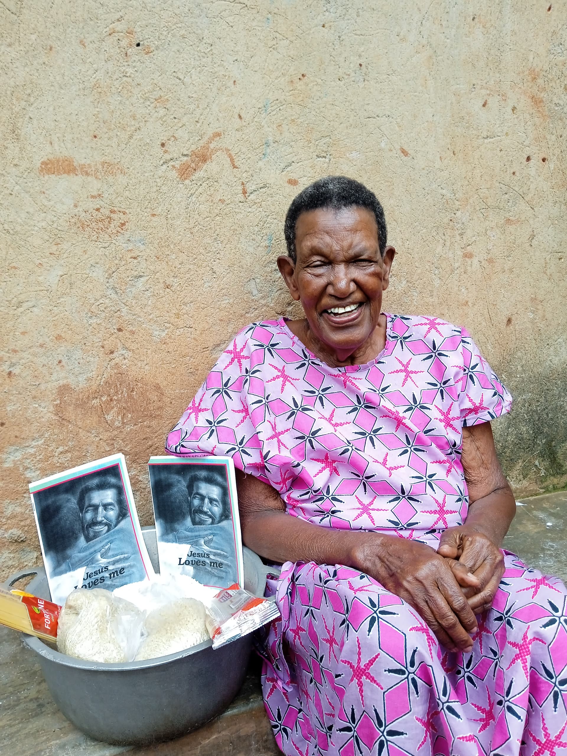 Woman in a pink floral dress sits laughing with a basin of food and photos on her lap.