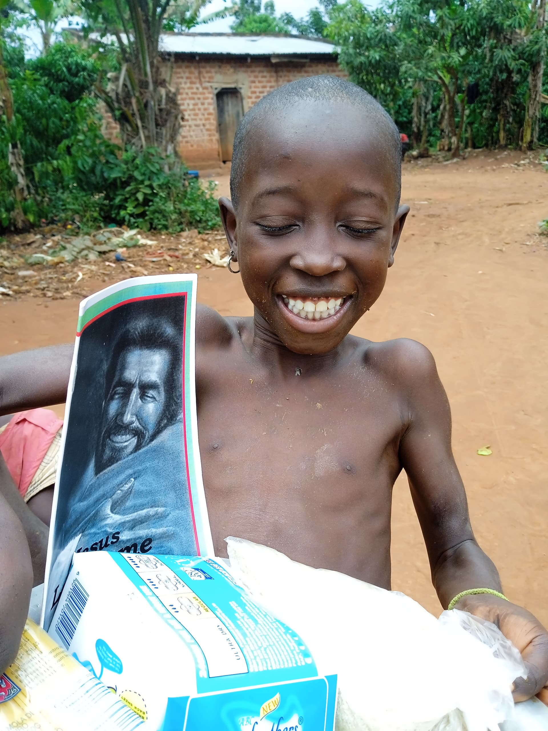 Boy smiles while holding a bag of food supplies with a photo booklet attached, dirt road behind him.