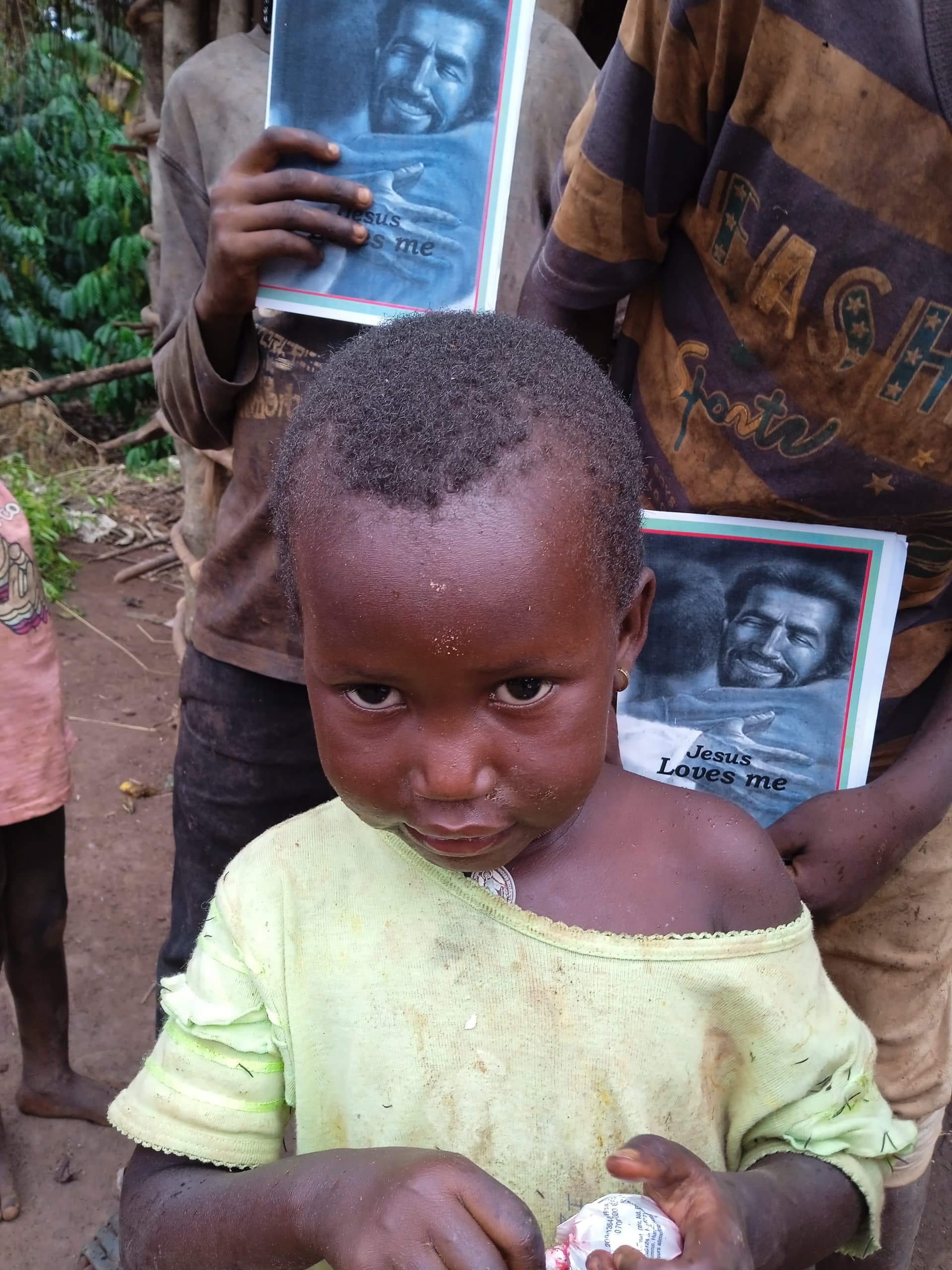 Young child in a yellow shirt stands with an adult holding a photo booklet near a mud hut.