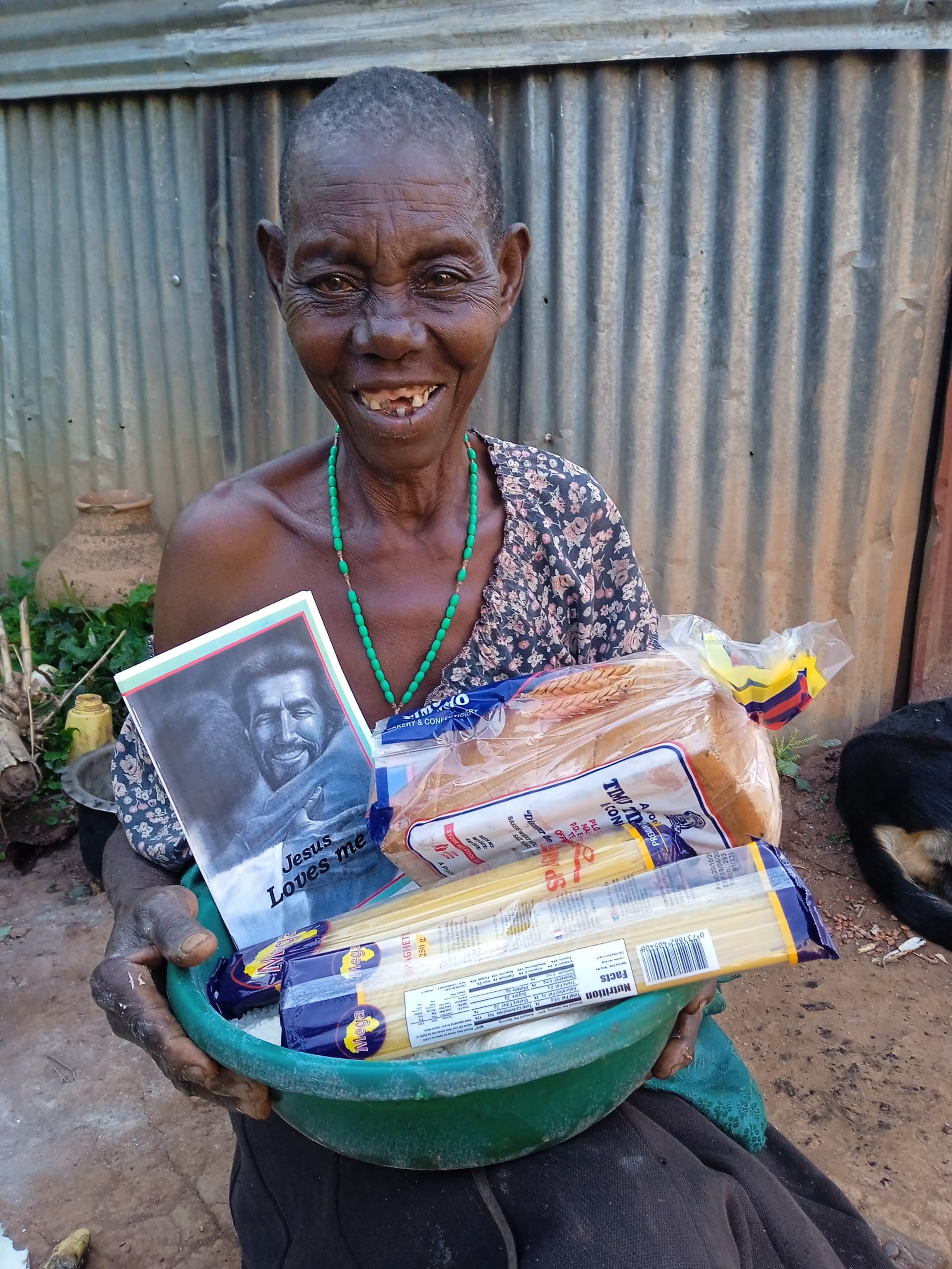 Elderly woman with a beaded necklace holds a green basin of pasta and cereal with a photo booklet.