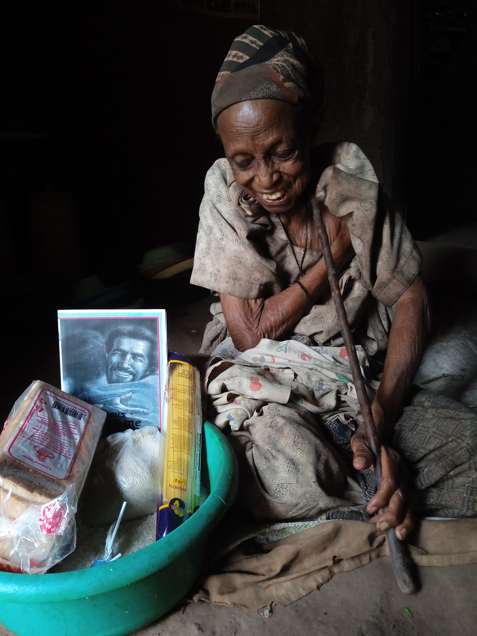 Elderly woman sits under a thatched shelter holding a basin of food supplies and a photo.