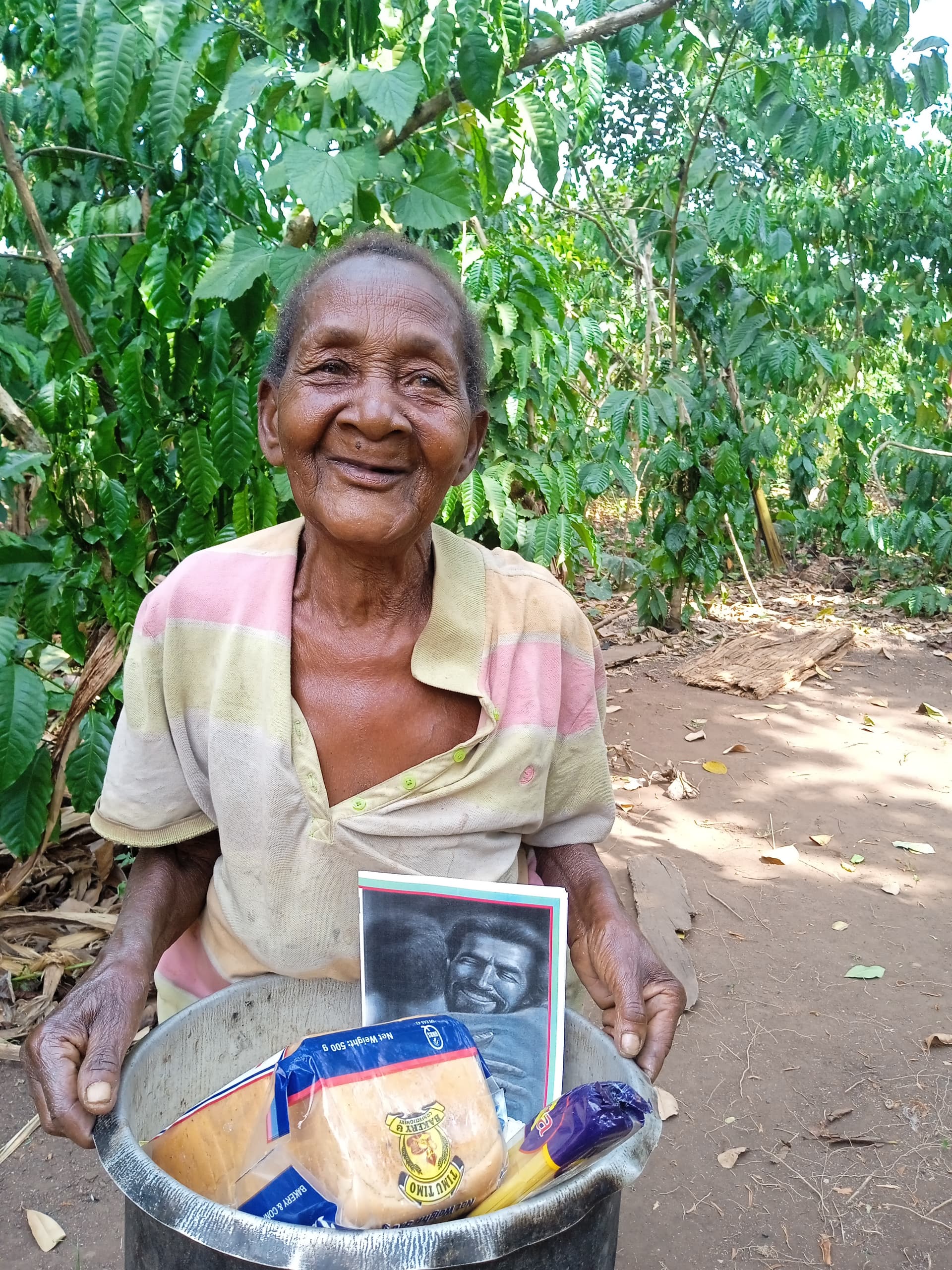 Elderly woman smiles outdoors while holding a basin of food supplies and a photo booklet.