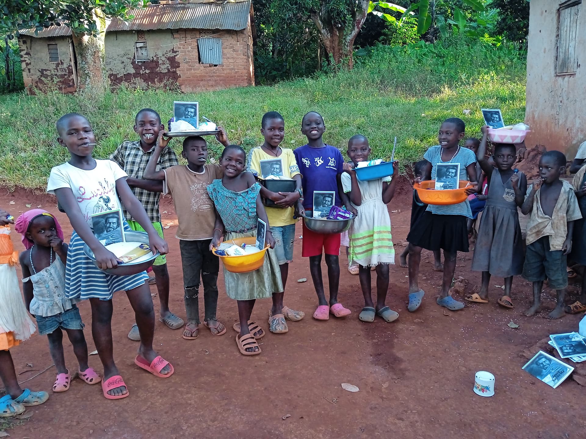Group of children stand outdoors near mud homes, some holding photo booklets from JFM.