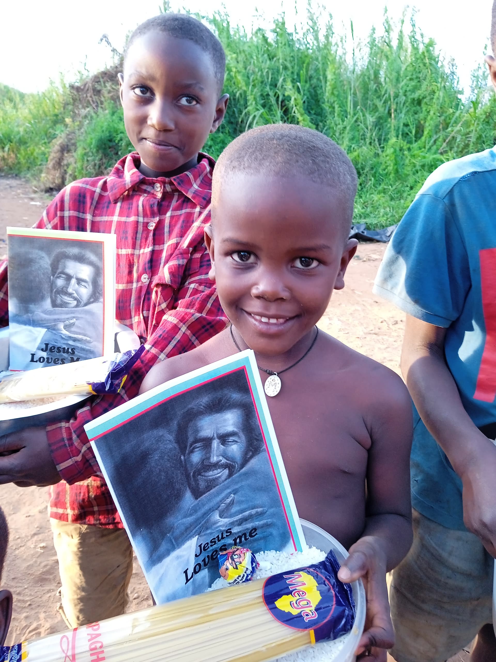 Boy in a plaid shirt holds a basin of rice, pasta, and a photo booklet, other children nearby.