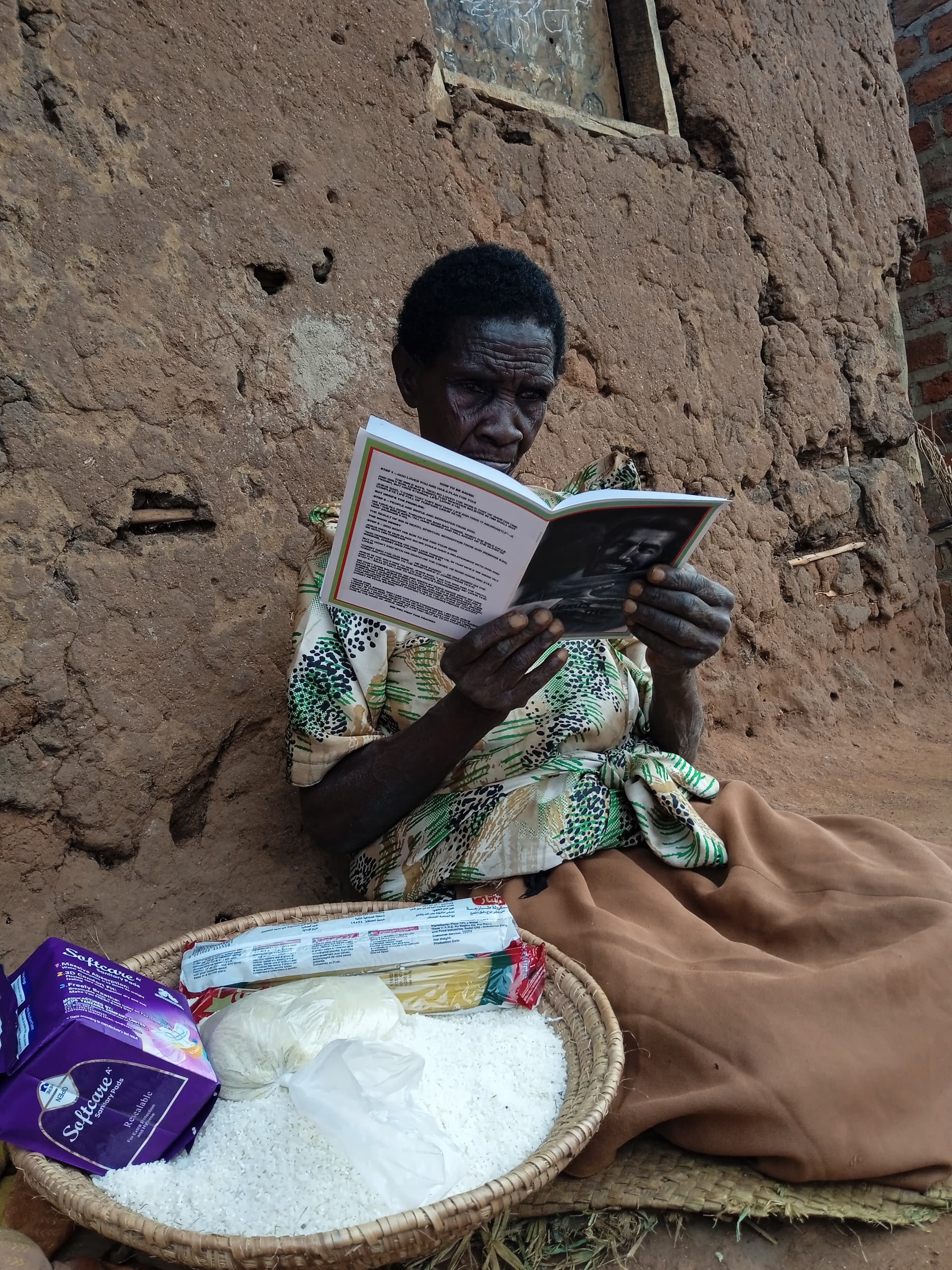 Woman sitting outdoors reading a booklet beside a basket holding a bag of rice.