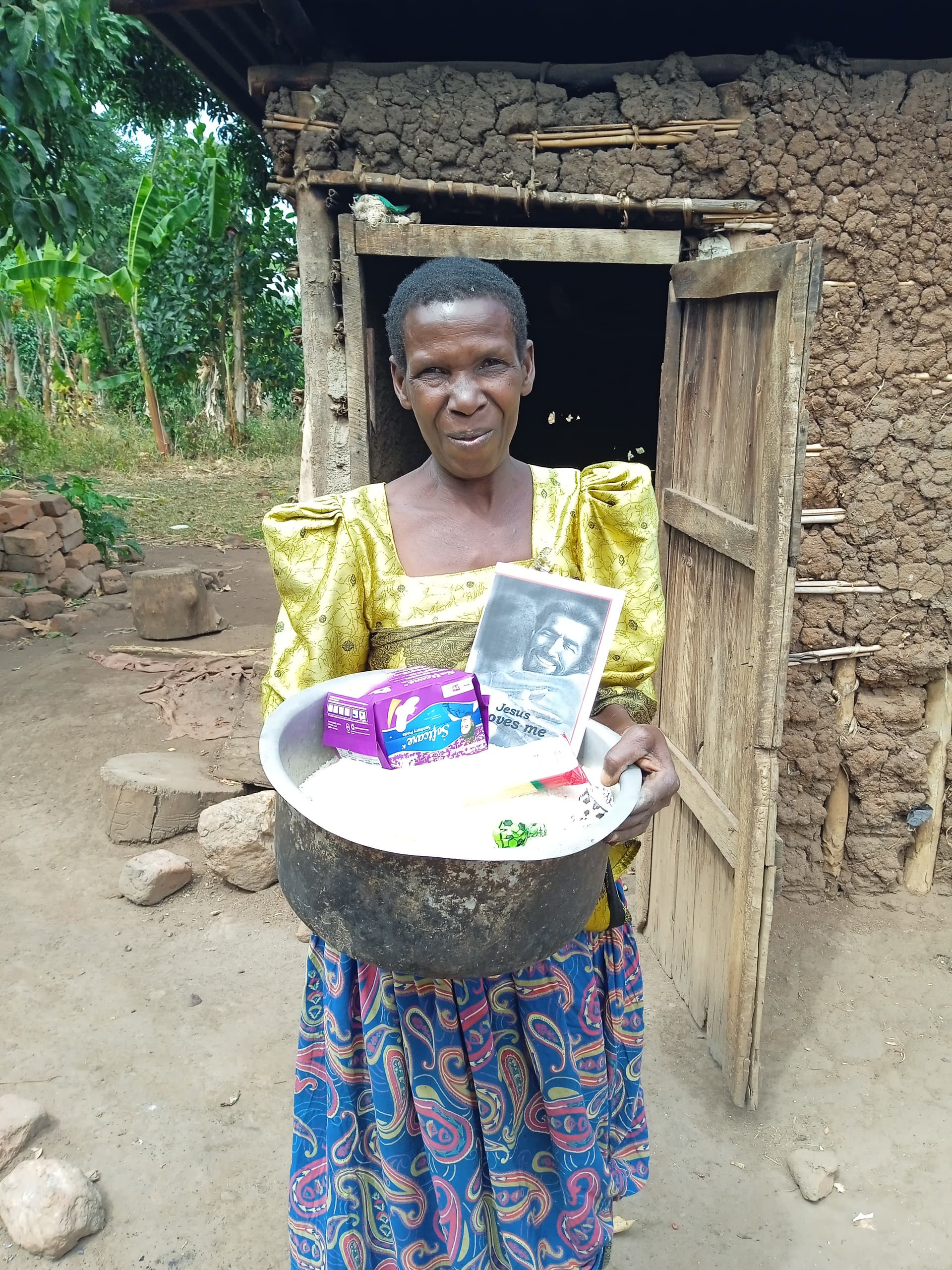 Woman standing in a doorway holding a basin of rice, soap, and a JFM photo booklet.