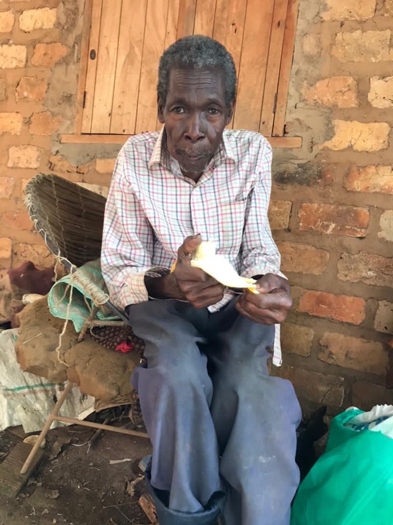 Elderly man Nkuutu Shaban seated indoors in a checkered shirt, reading a gospel tract.