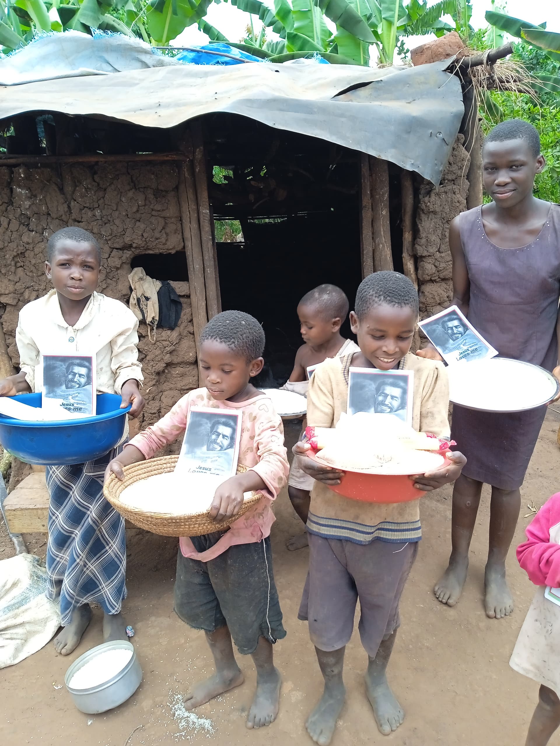 Group of children standing outside a thatched shelter, holding food basins and tract booklets.