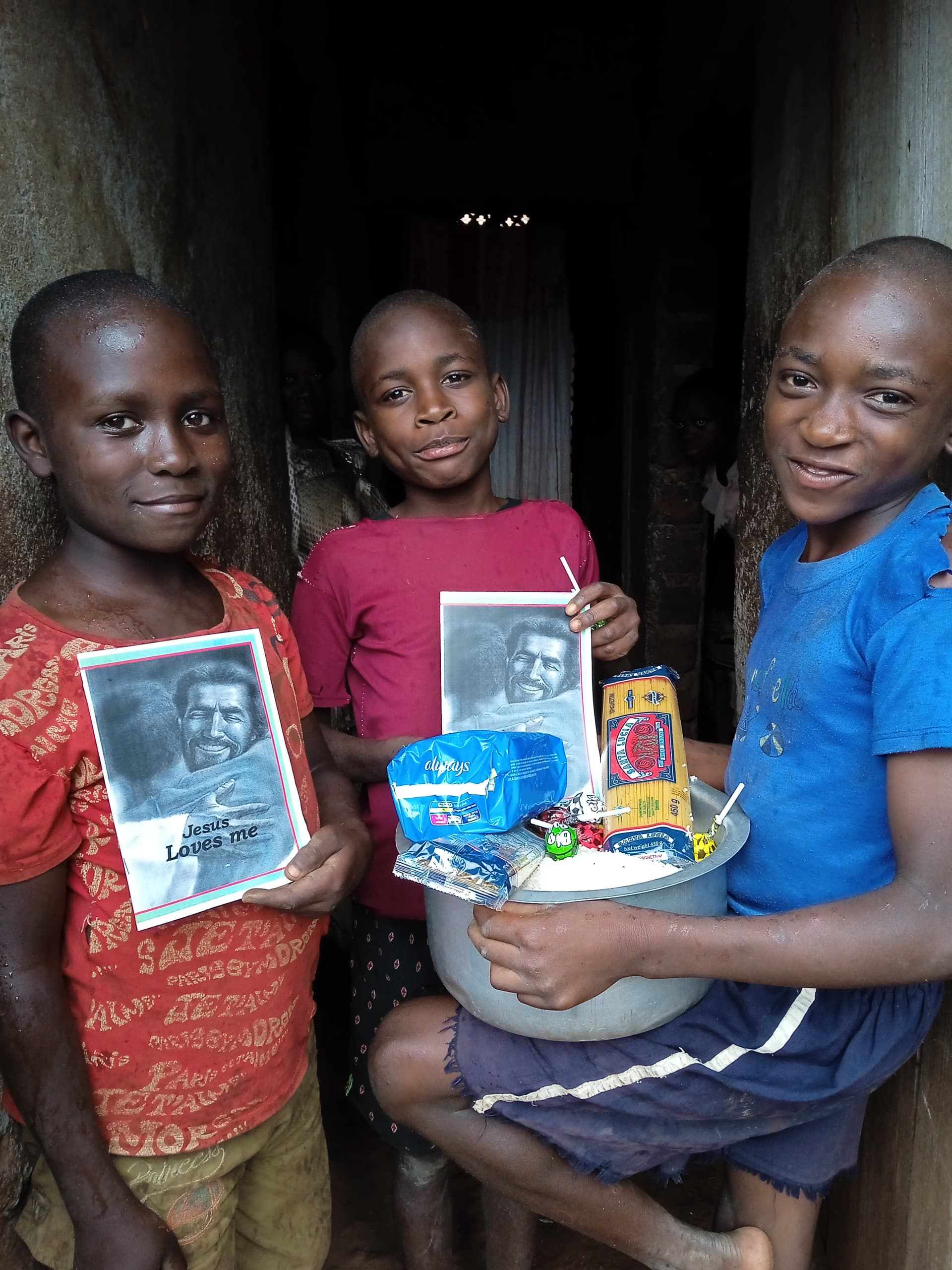 Two children standing together in a doorway, holding a gospel tract booklet and food basin.
