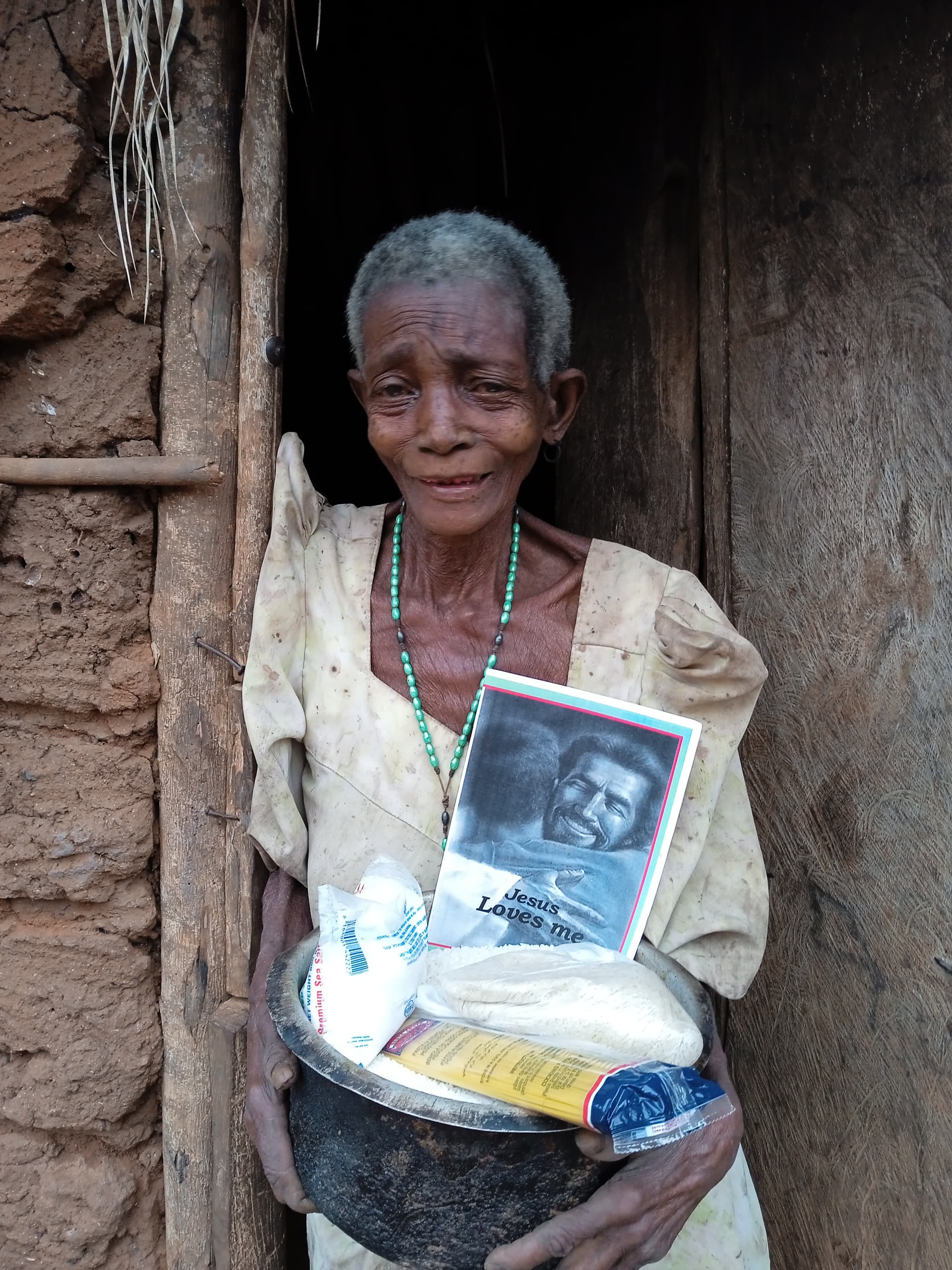 Elderly woman standing in a doorway, holding a gospel tract booklet and metal basin of food.