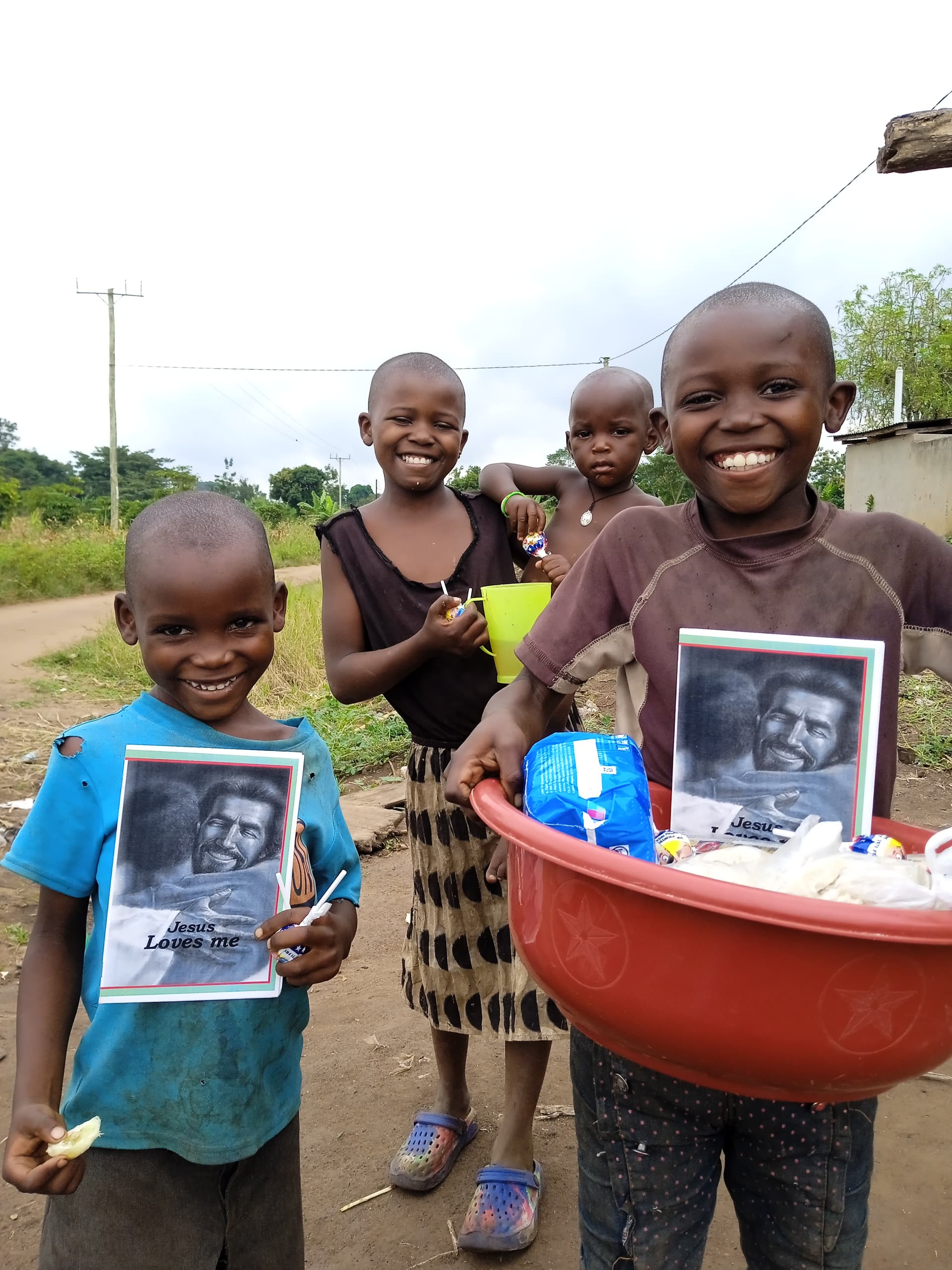 Three children standing on a dirt road, one holding a tract booklet and red basin of food.