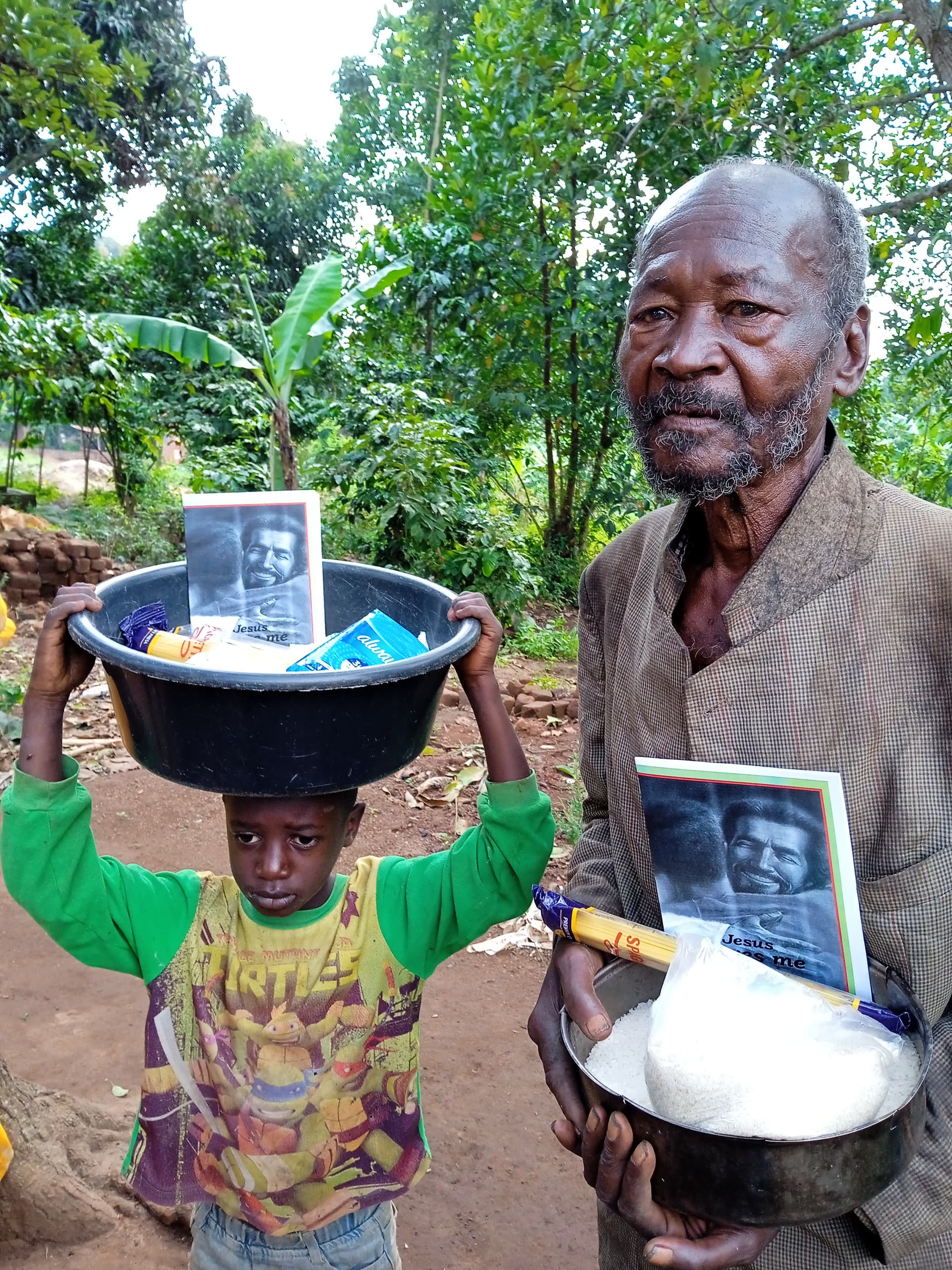 Elderly man standing with a boy carrying a basin of food and tract booklet on his head.