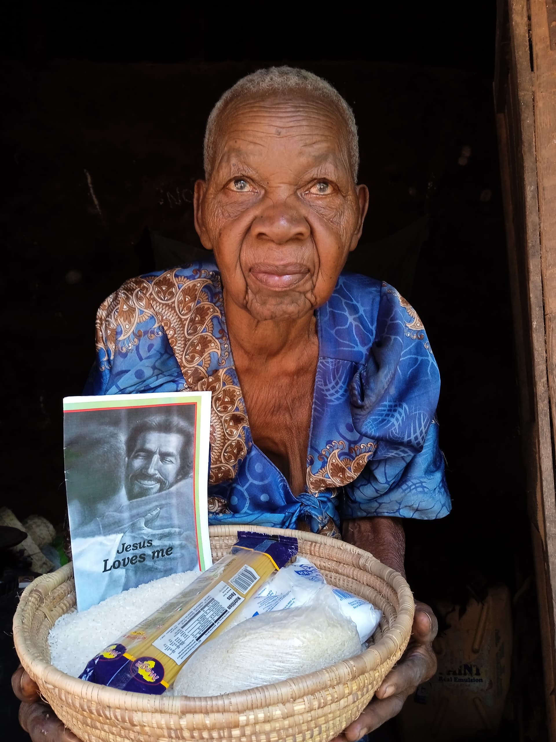 Elderly woman standing in a doorway holding a gospel tract booklet and basket of food.
