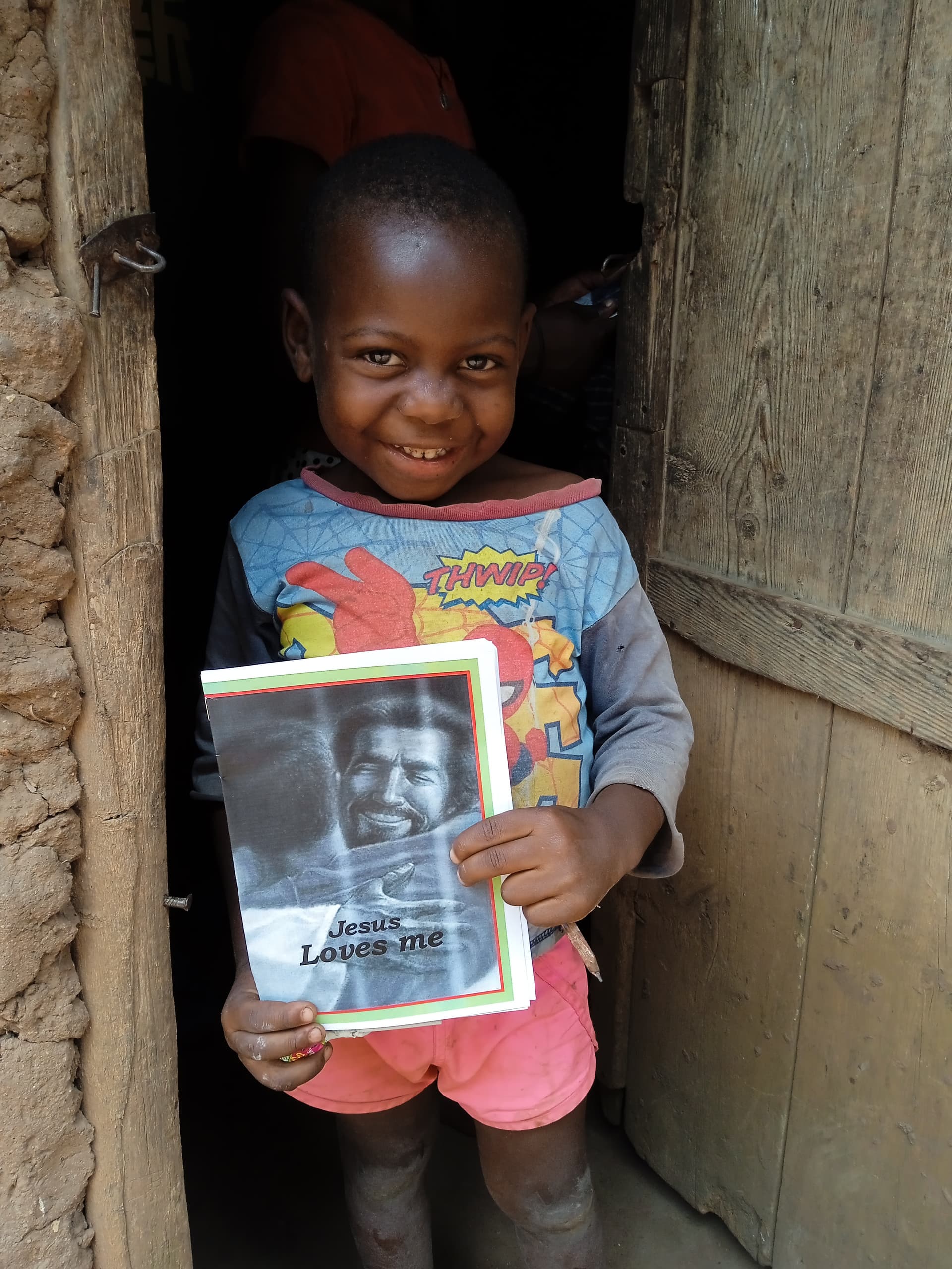 Young boy smiling in the doorway of a mud home, holding a gospel tract booklet.
