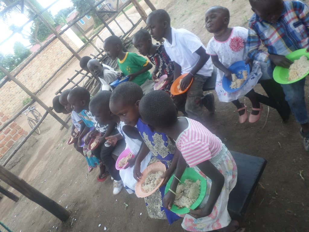 Row of children seated together outdoors near a fence, eating from plates and bowls.
