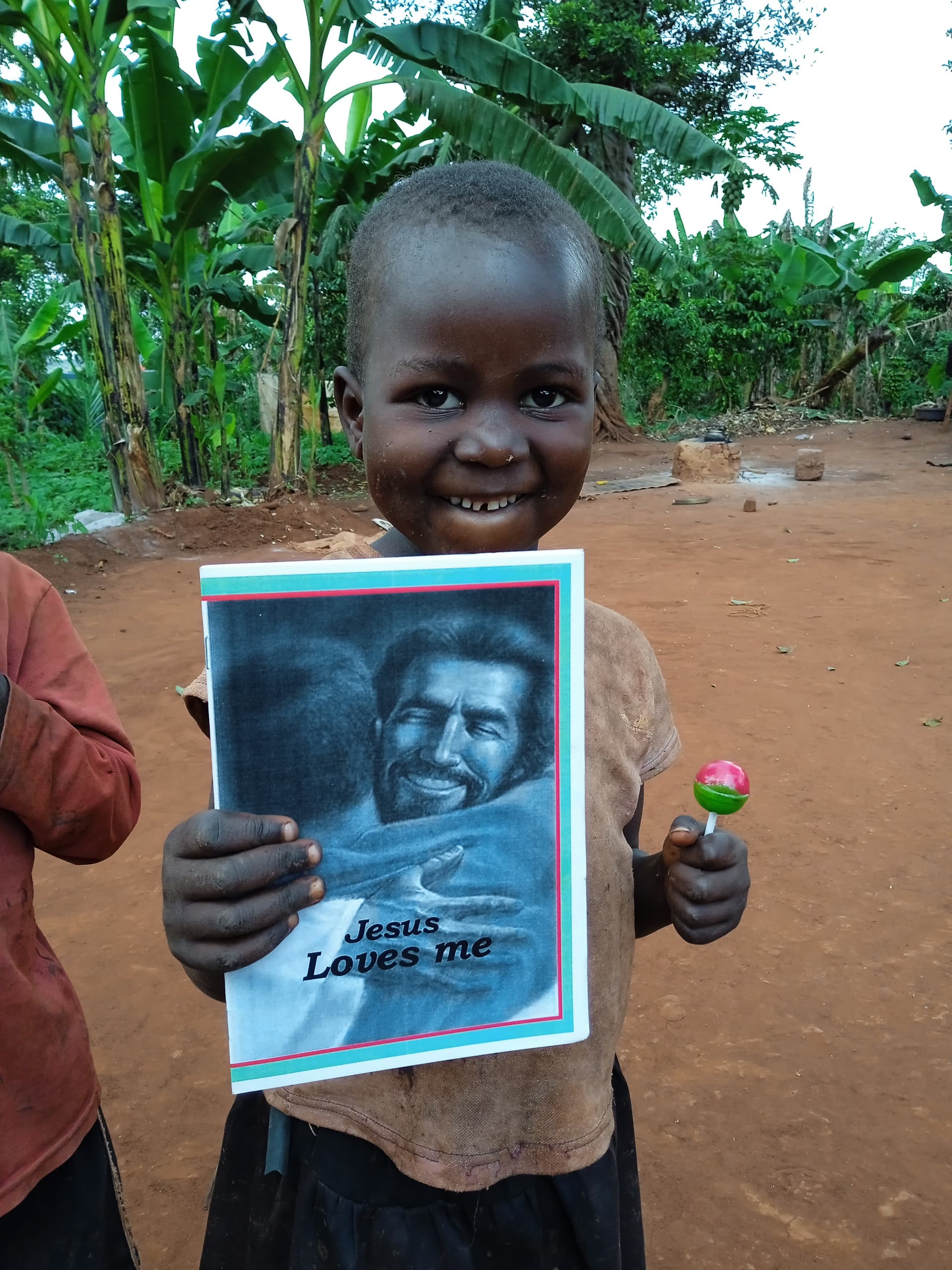Child standing outdoors near banana plants, holding a Jesus Loves Me gospel tract and small toy.
