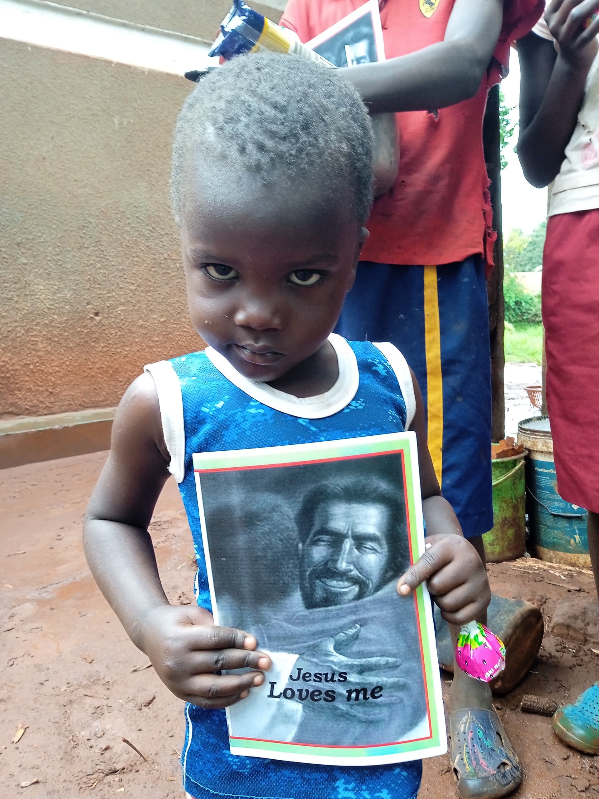 Child in a blue tank top standing outdoors holding a Jesus Loves Me gospel tract.