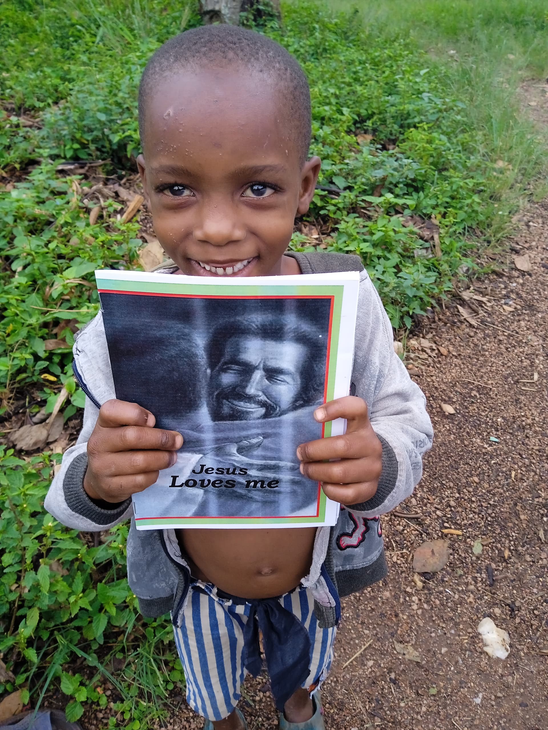 Boy standing outdoors on a dirt path, smiling and holding a Jesus Loves Me gospel tract.