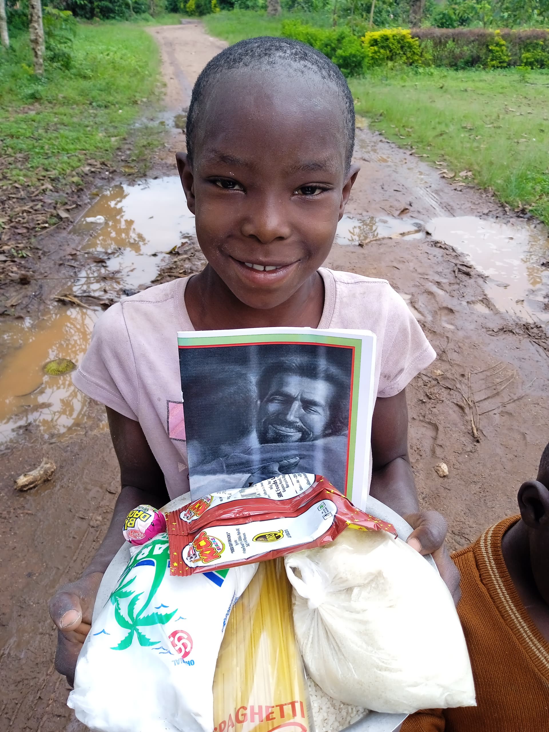 Girl standing on a dirt road holding a photo card and packages of food supplies.